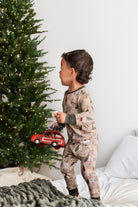 A toddler stands on a bed wearing a beige holiday-themed pajama set with a retro light print, holding a red toy car next to a decorated Christmas tree.