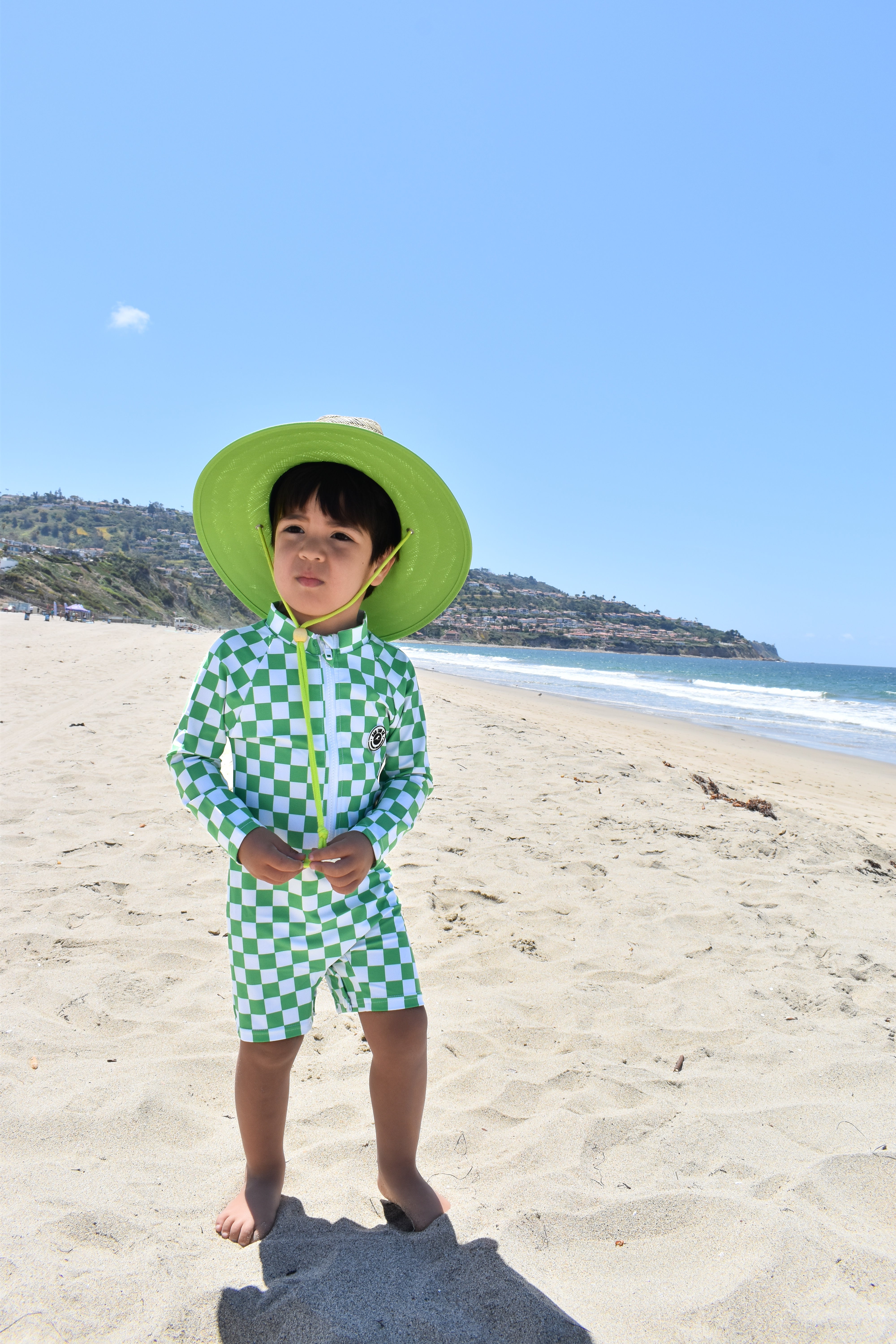Child in green checkered swimsuit and hat standing on a sandy beach with ocean and clear sky in the background.