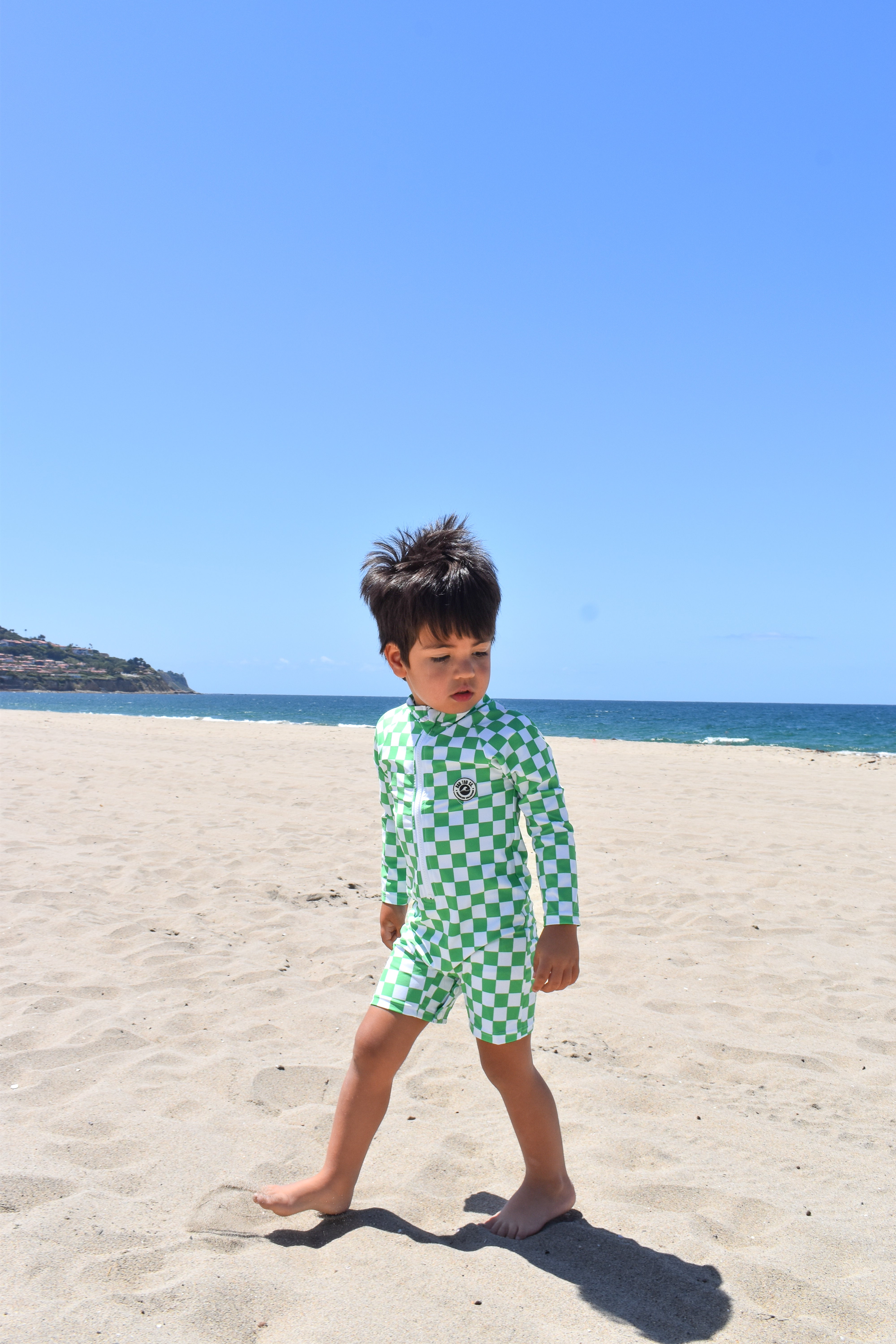 Child in a green checkered swimsuit standing on a sandy beach with clear blue sky.