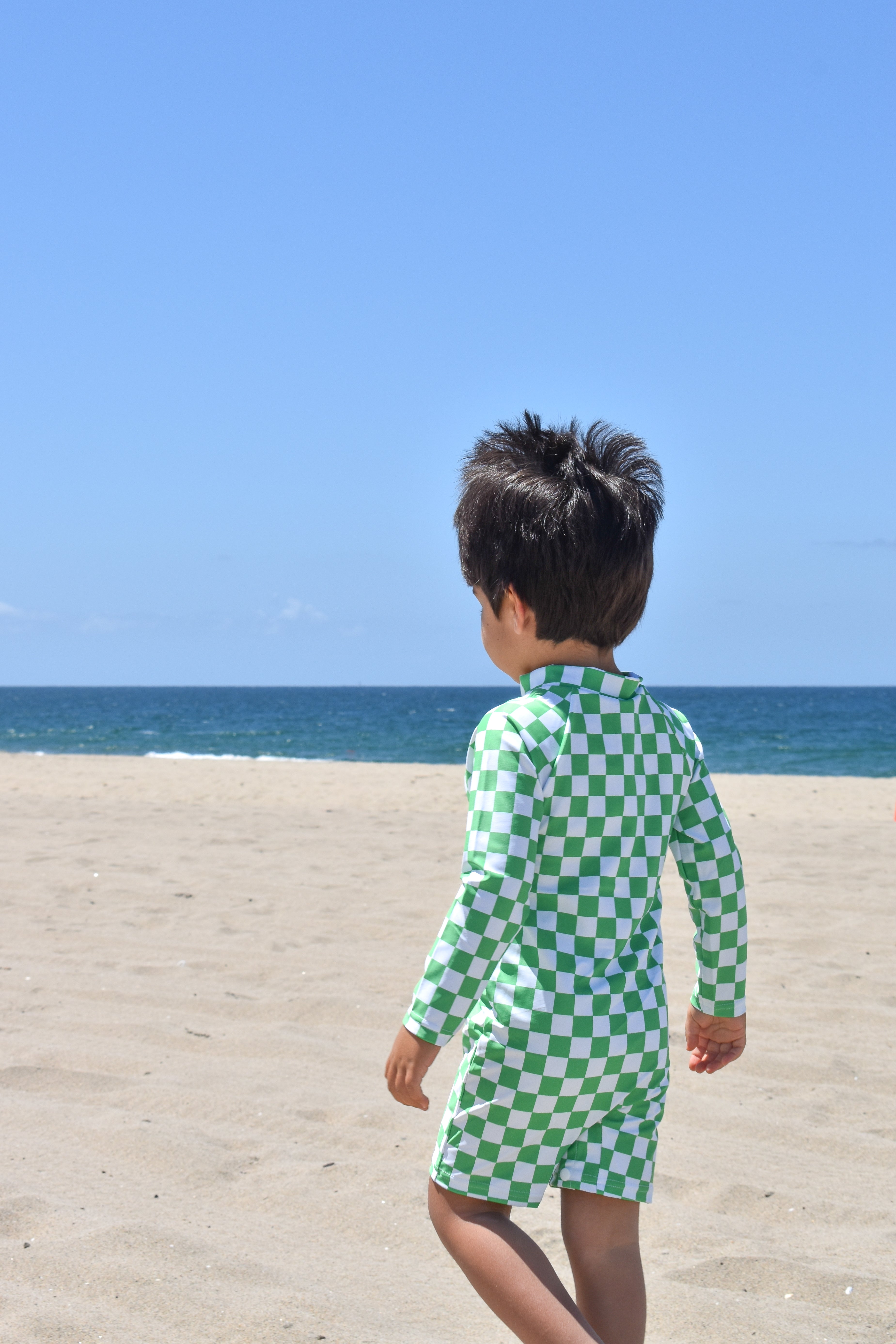 Child wearing a green checkered swimsuit standing on a sandy beach with ocean in the background
