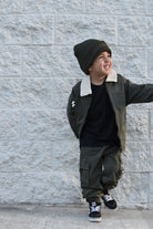 A young boy wearing a dark olive green jacket with a white shearling collar, matching cargo pants, a black t-shirt, and a dark knit beanie, posing against a light gray textured brick wall.