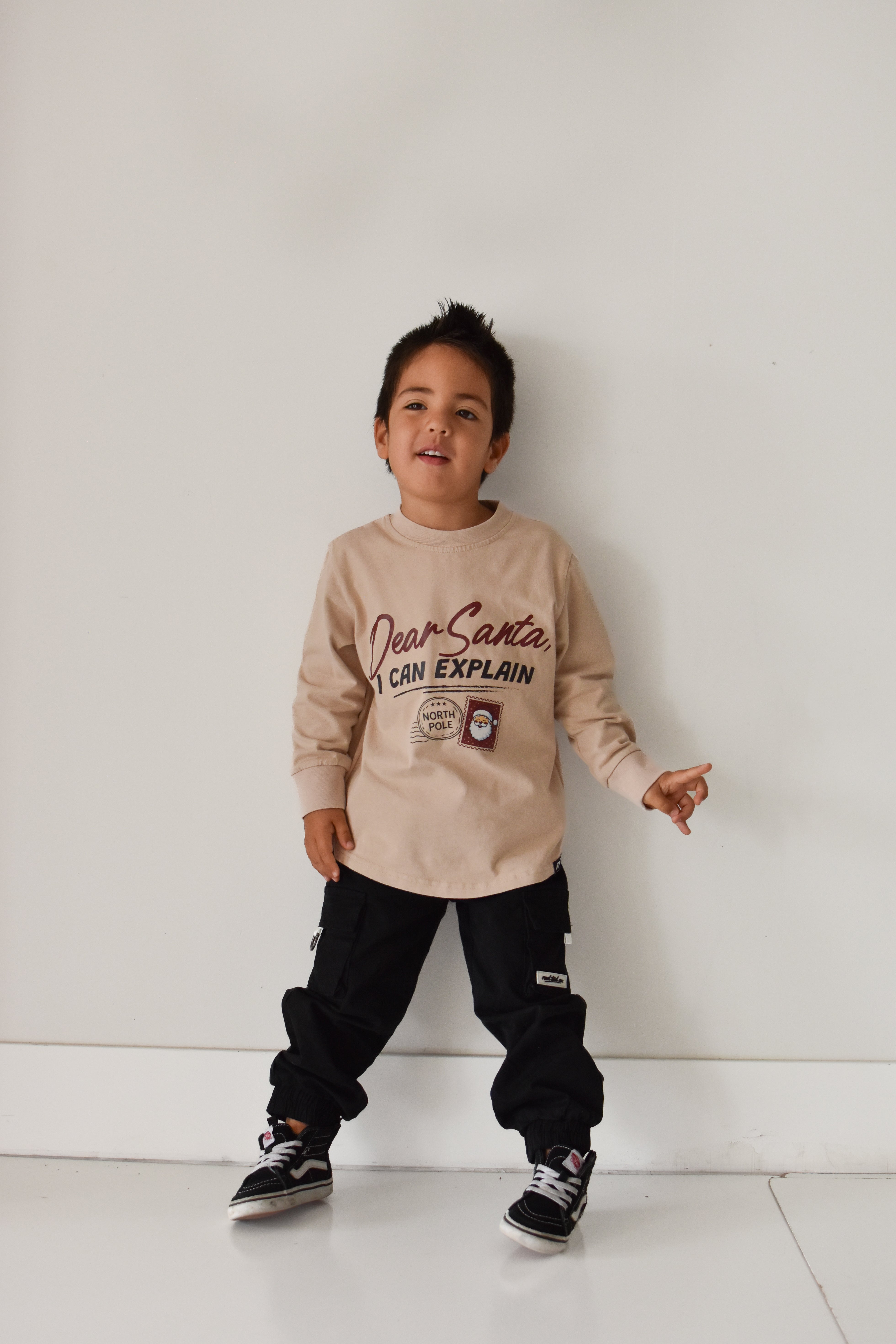A young boy wearing a beige long-sleeve graphic sweatshirt and black cargo pants, standing against a plain white wall.