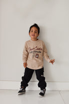 A young boy wearing a beige long-sleeve graphic sweatshirt and black cargo pants, standing against a plain white wall.