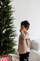 A young boy wearing a beige long-sleeve graphic t-shirt and black pants stands next to a decorated Christmas tree in a bright room with a white bed in the background.
