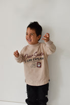 A young boy smiling and posing in a beige long-sleeve graphic sweatshirt that reads 'Dear Santa I CAN EXPLAIN' with a Santa patch graphic, paired with black pants, set against a plain white wall.