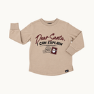 A beige long-sleeve shirt for kids, featuring a graphic print on the front that reads 'Dear Santa, I CAN EXPLAIN' with a postmark design and a small Santa illustration. The shirt is shown against a plain off-white background.