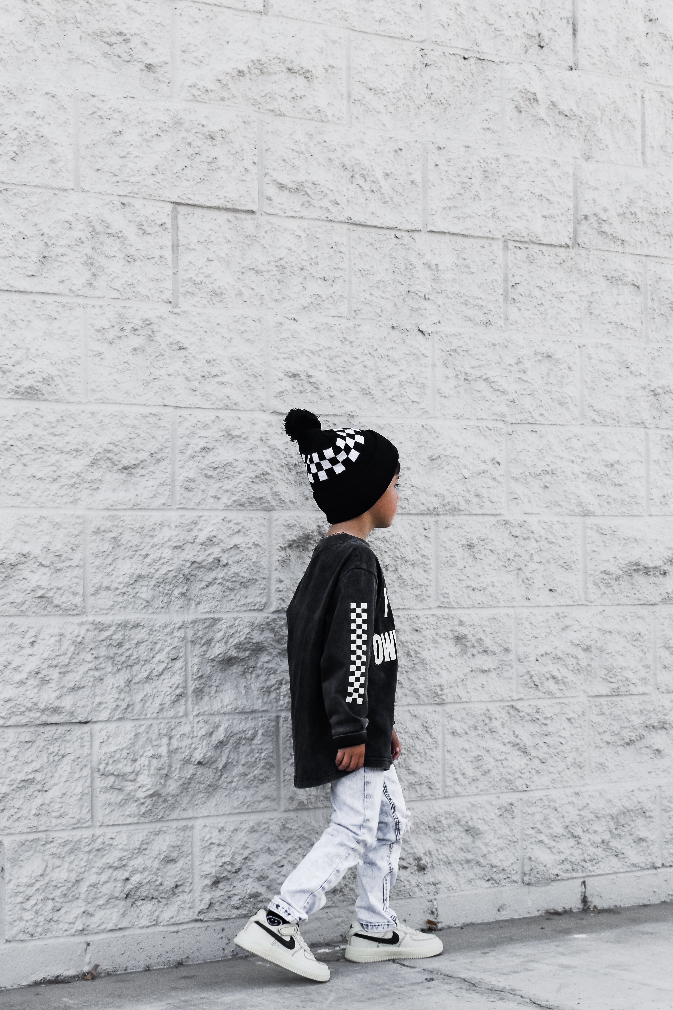 Child wearing a black long-sleeve shirt with white text, white pants, and a checkered beanie against a light gray brick wall.