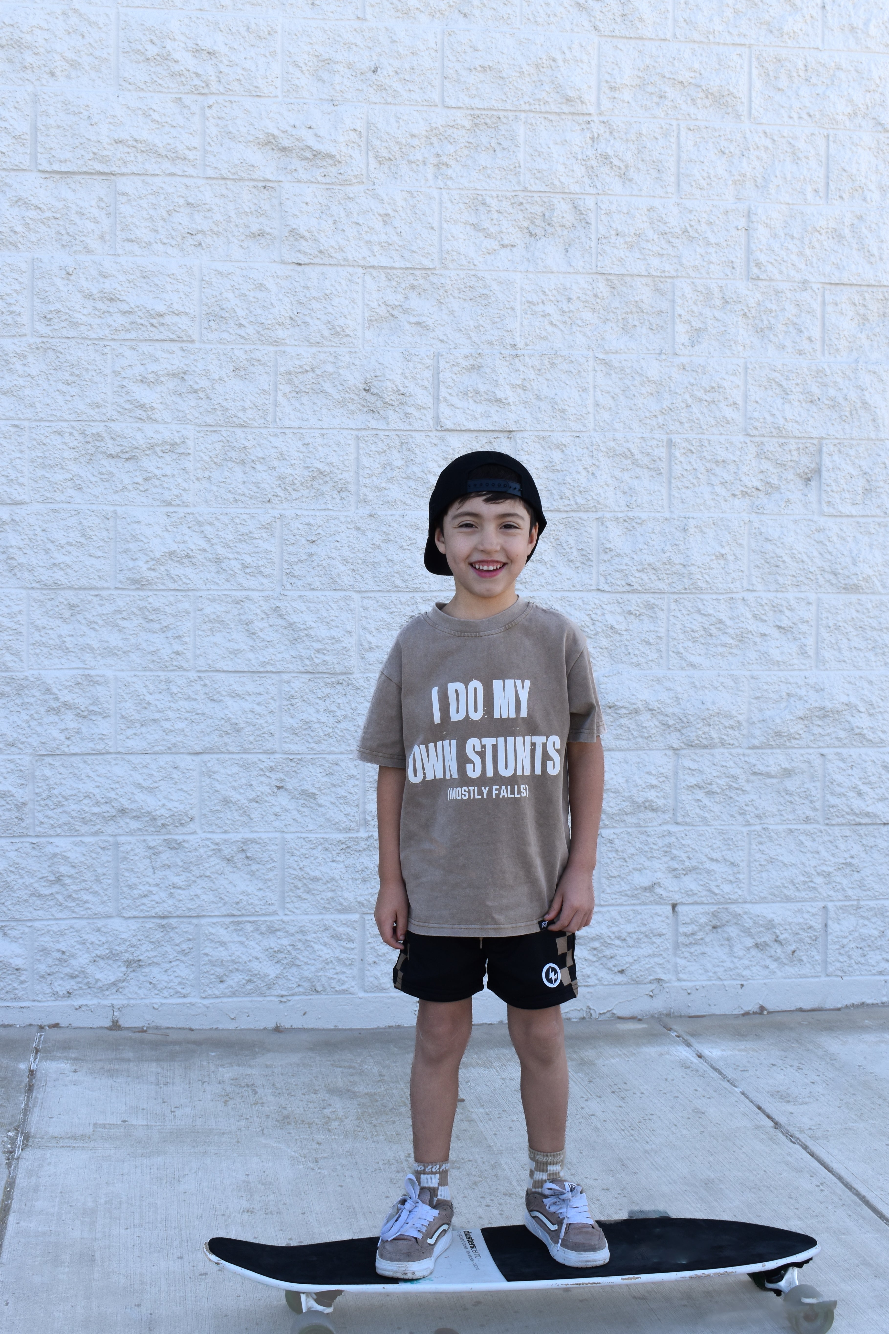 Child standing on a skateboard wearing a t-shirt with text, against a light stone wall.