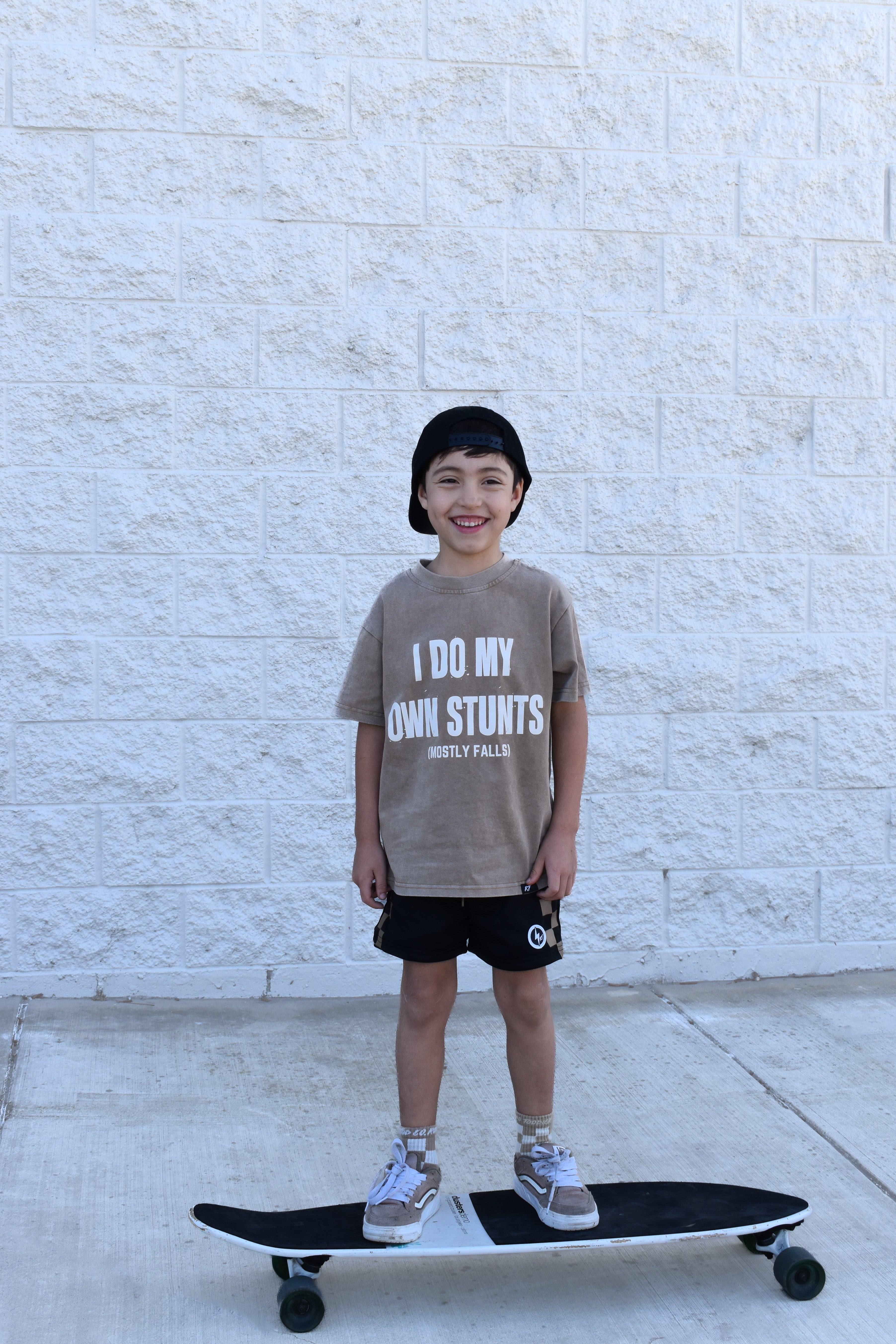 Child standing on a skateboard wearing a t-shirt with text, against a light stone wall.