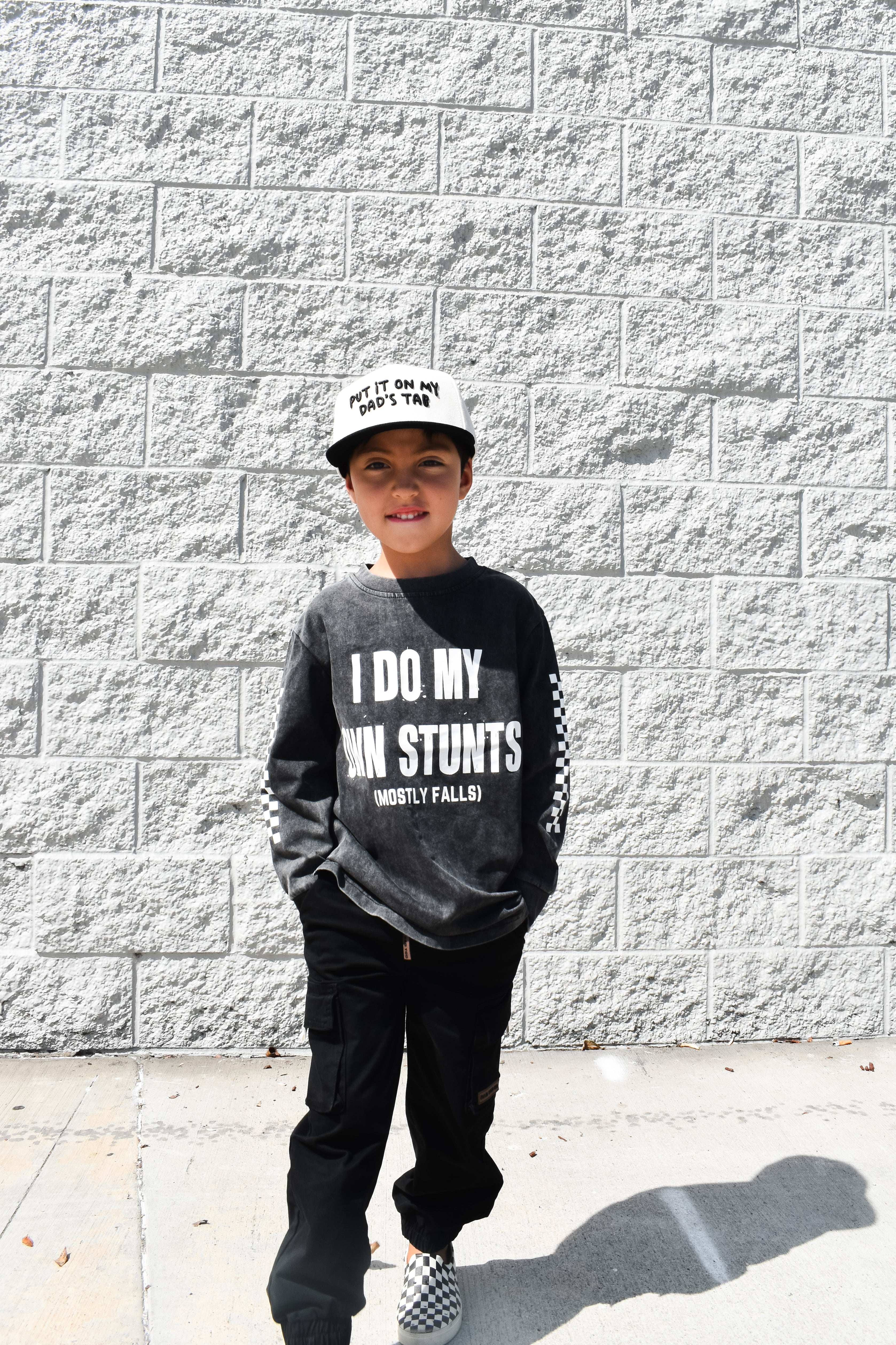 Child wearing a black sweatshirt with text, standing against a gray brick wall.