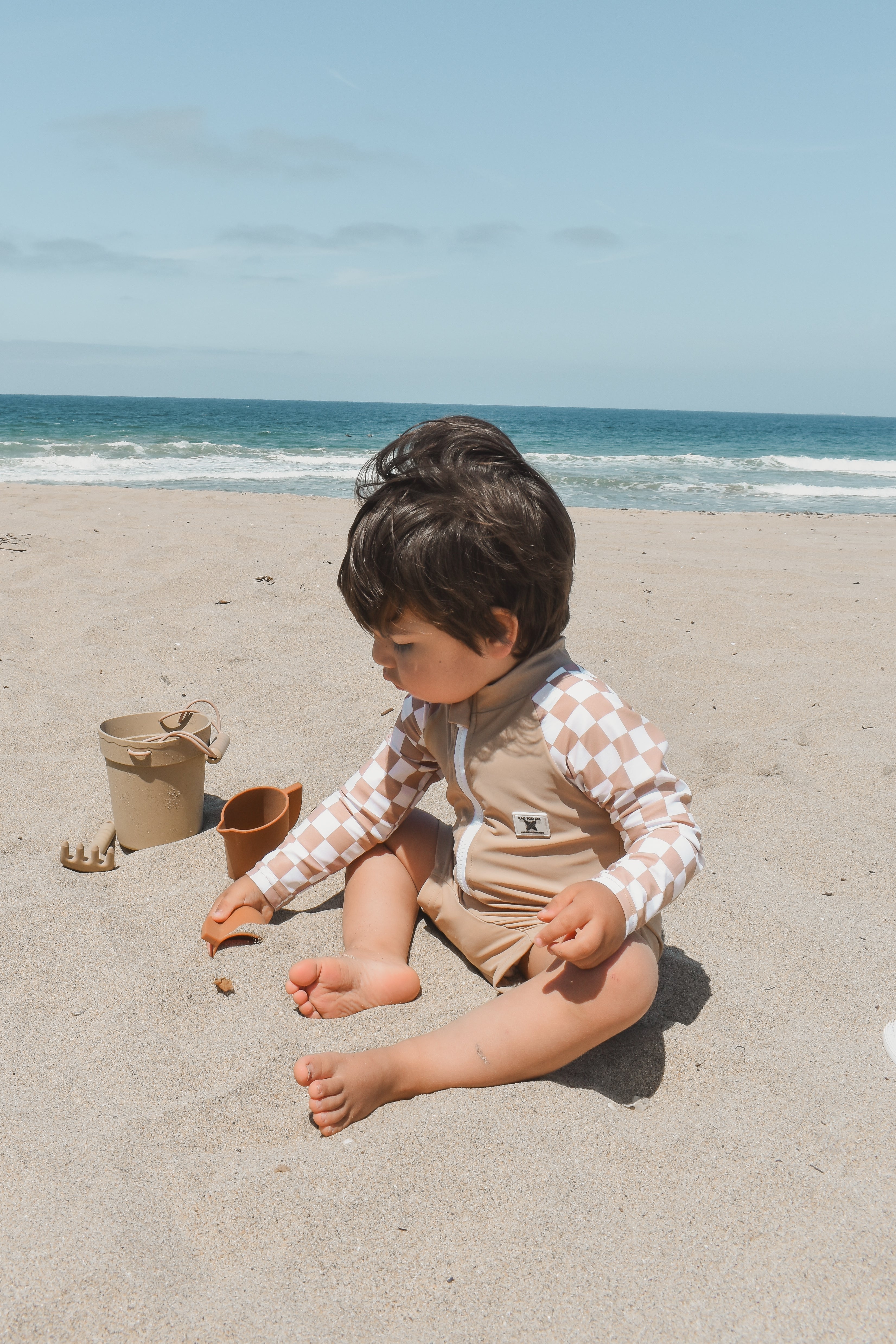 Toddler wearing the Dune Checkers one-piece rashguard with beige body and white checker sleeves, playing in the sand.