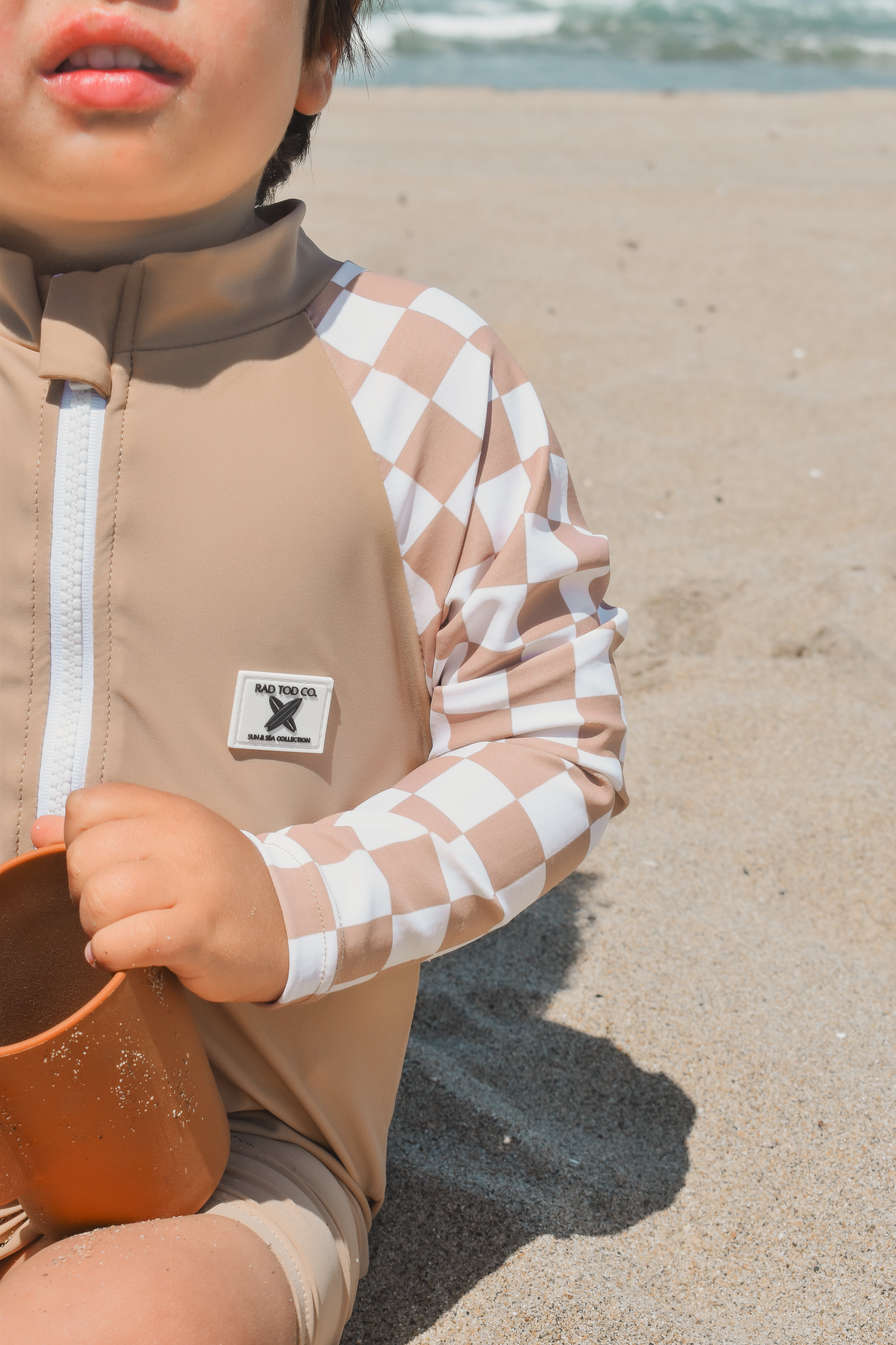 Toddler wearing the Dune Checkers one-piece rashguard with beige body and white checker sleeves, sitting on the beach.