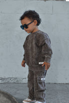 A toddler wearing a matching washed dark brown sweatshirt and cargo pants set, styled with black sunglasses and white sneakers, standing against a light gray concrete wall.