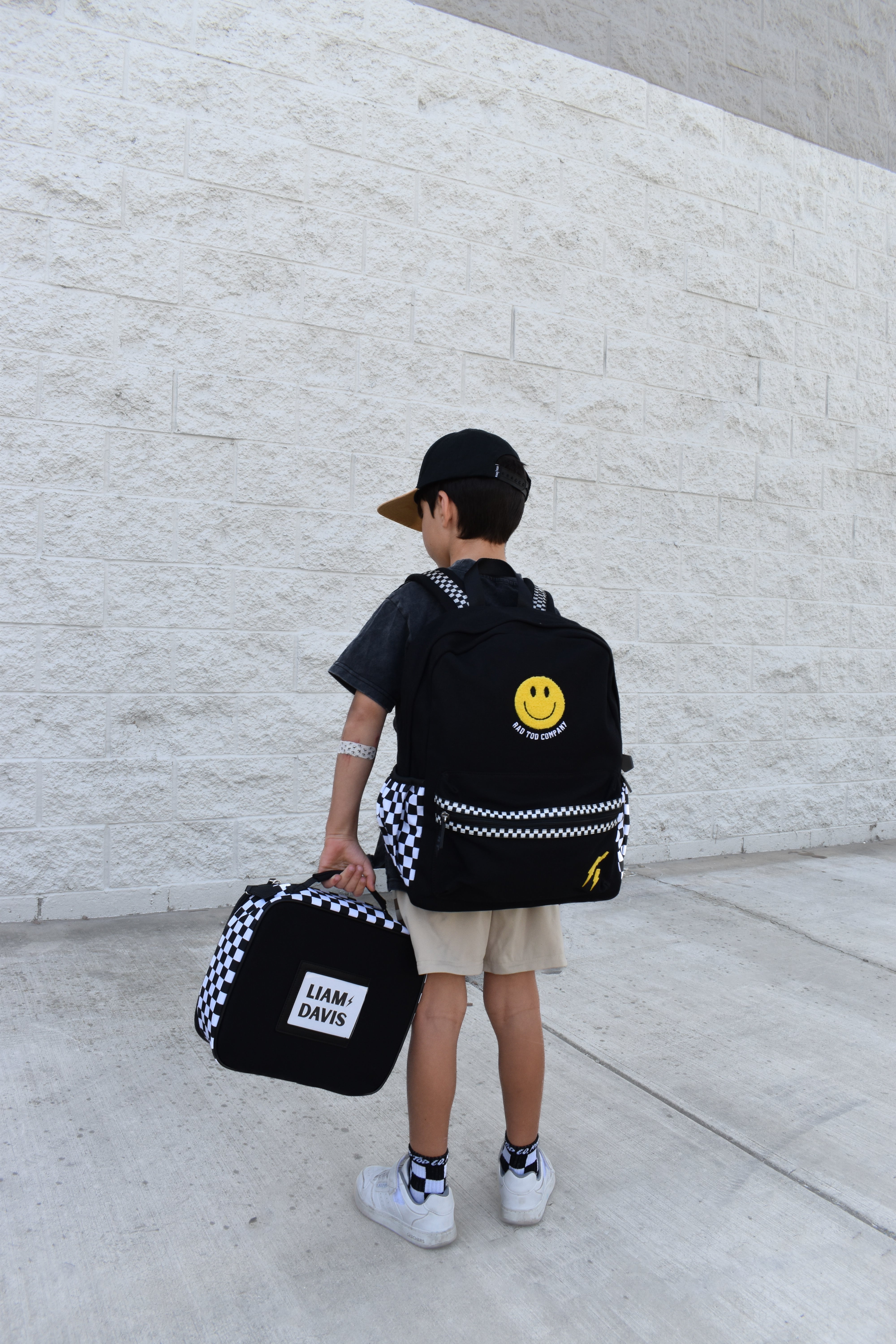 Child holding a black backpack with a smiley face design and a smaller bag, standing against a light gray wall.