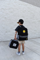 Child holding a black backpack with a smiley face design and a smaller bag, standing against a light gray wall.