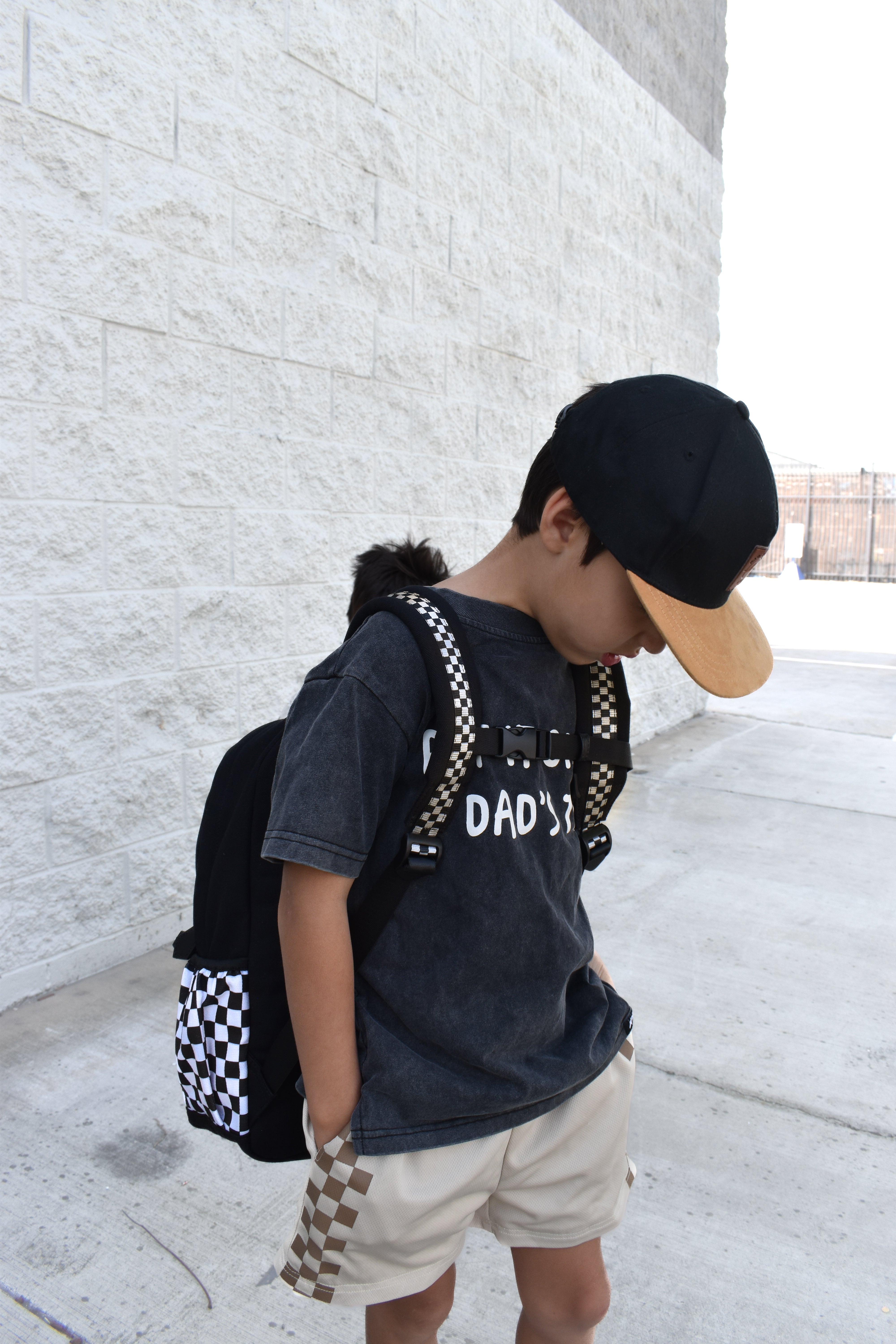 Child wearing a black t-shirt with text, beige shorts, and a checkered backpack against a light stone wall.