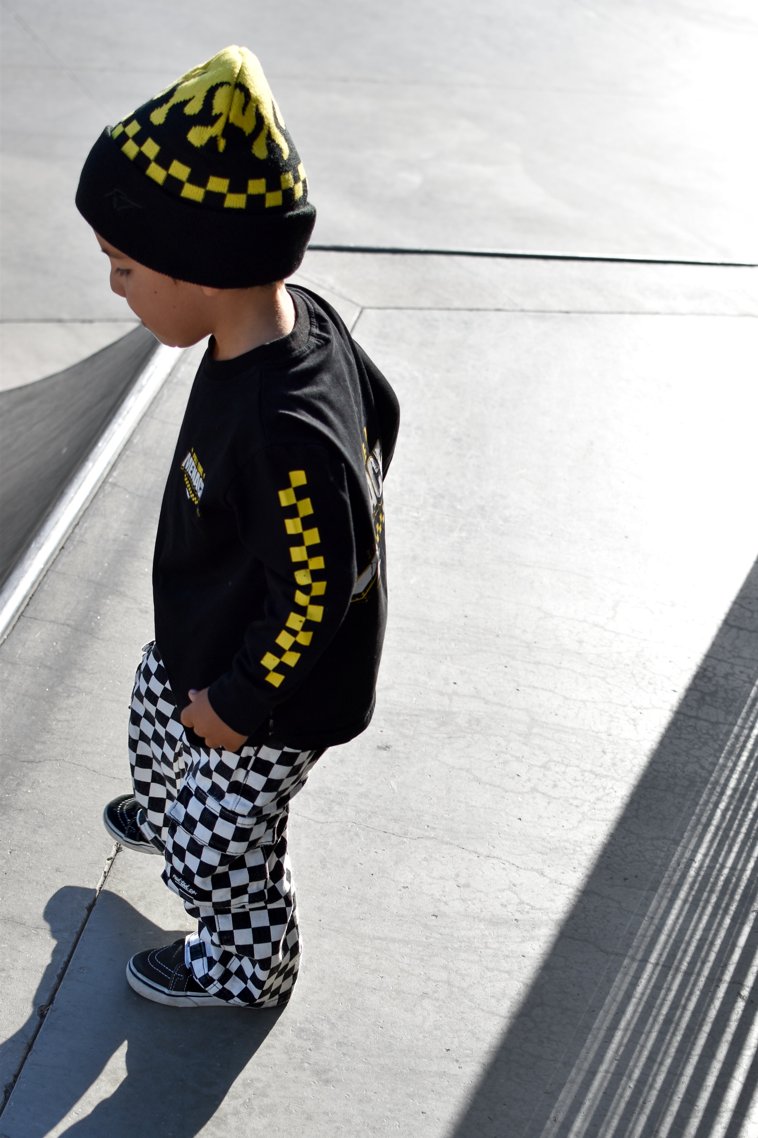 A toddler standing outdoors on a concrete surface, wearing a black beanie with yellow checkered accents, a black long-sleeve shirt with yellow checkered sleeve details, and black-and-white checkered pants.