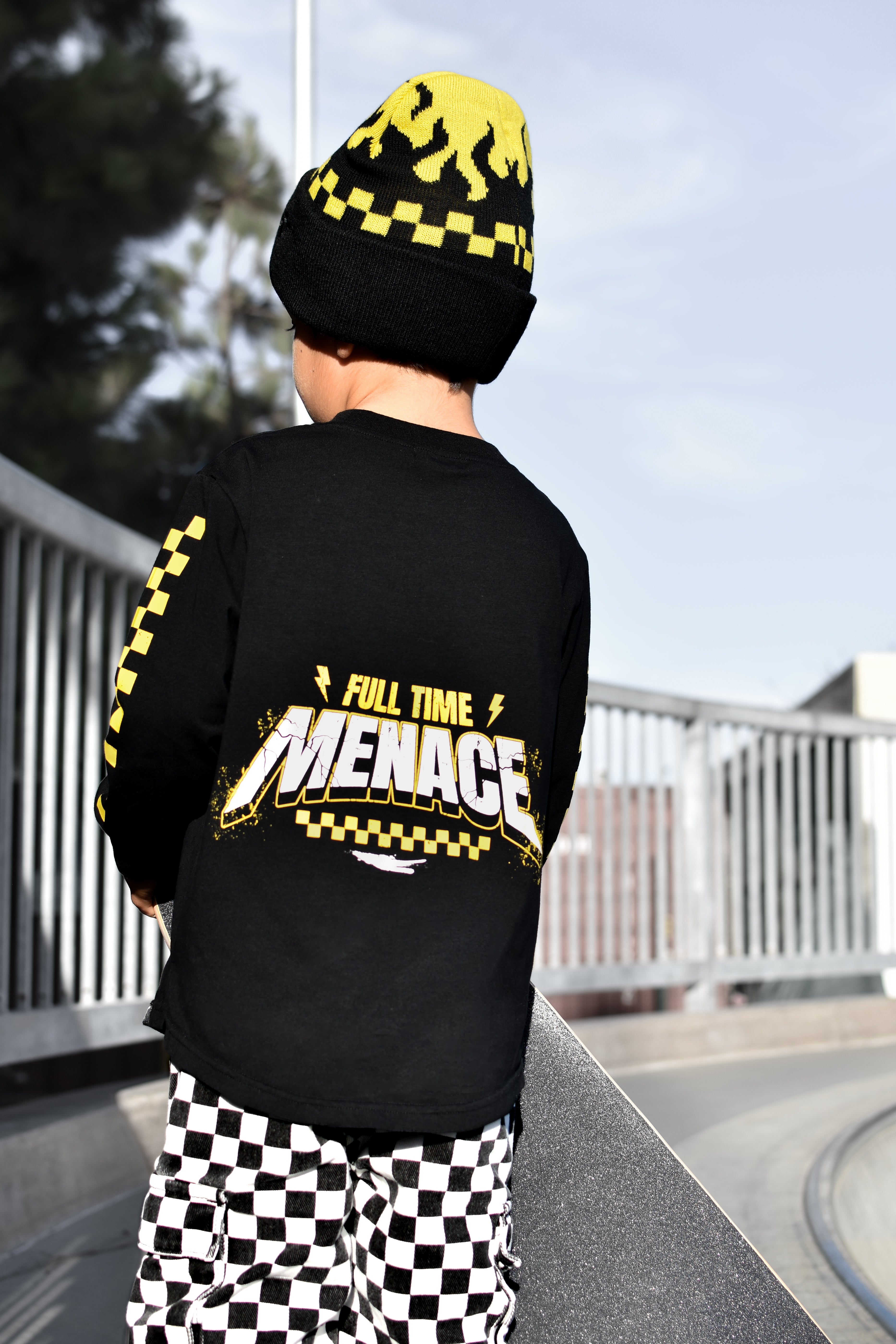 A child viewed from behind wearing a black long-sleeve t-shirt with a yellow and white 'Full Time Menace' graphic on the back, a matching black and yellow beanie, and black-and-white checkered pants, standing outdoors in a skatepark.