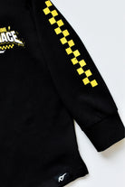 Close-up of a black long-sleeve shirt with a yellow and black checkered pattern down one sleeve and a partial graphic on the chest, shown on a light gray background.