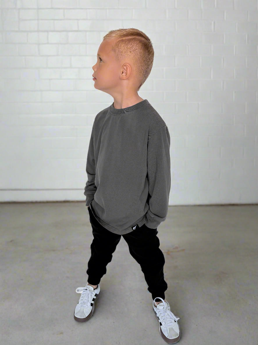 Child wearing a gray sweatshirt and black pants standing against a white brick wall.