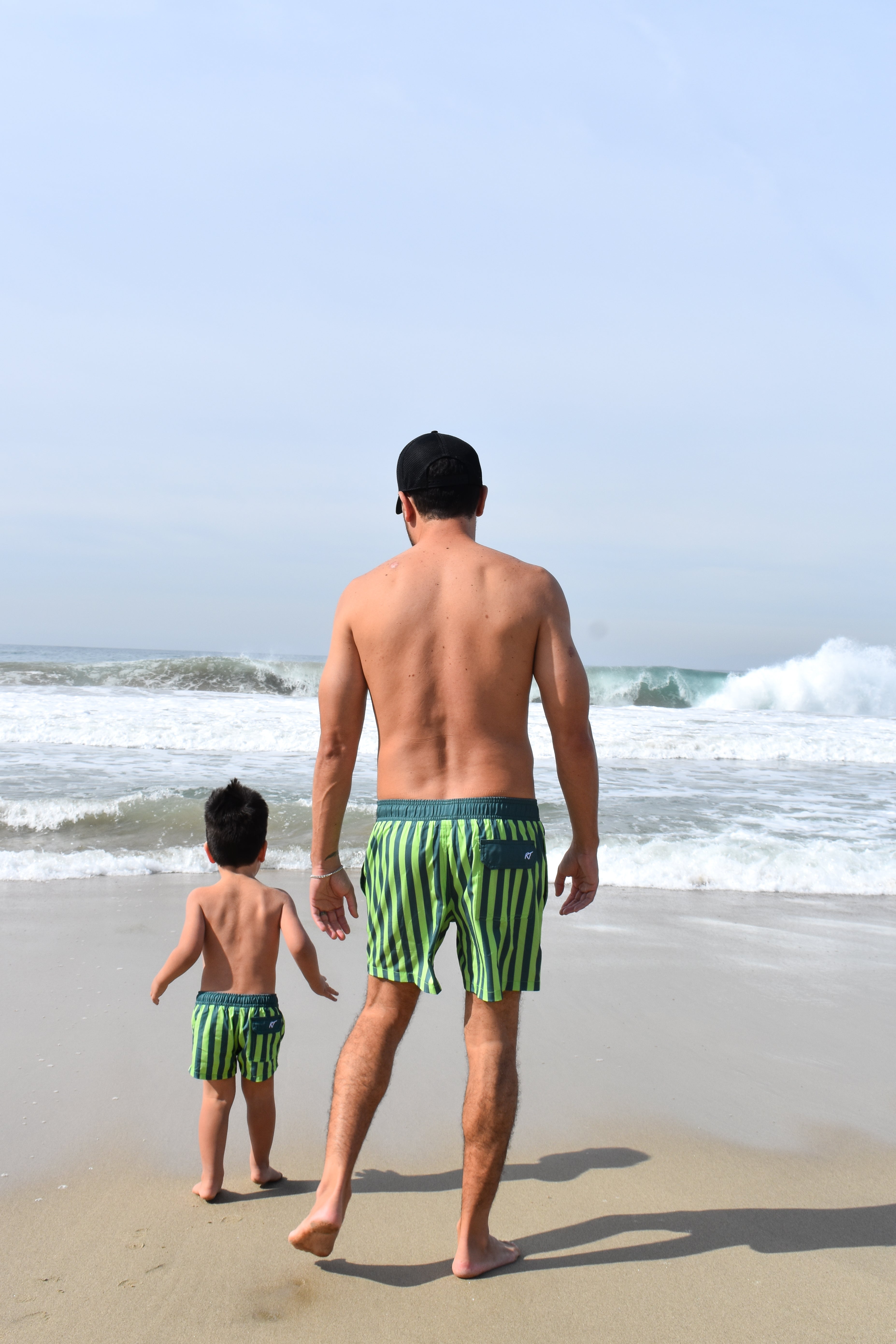 Man and child walking on a beach wearing matching lime green and dark green striped swim trunks by rad toddler