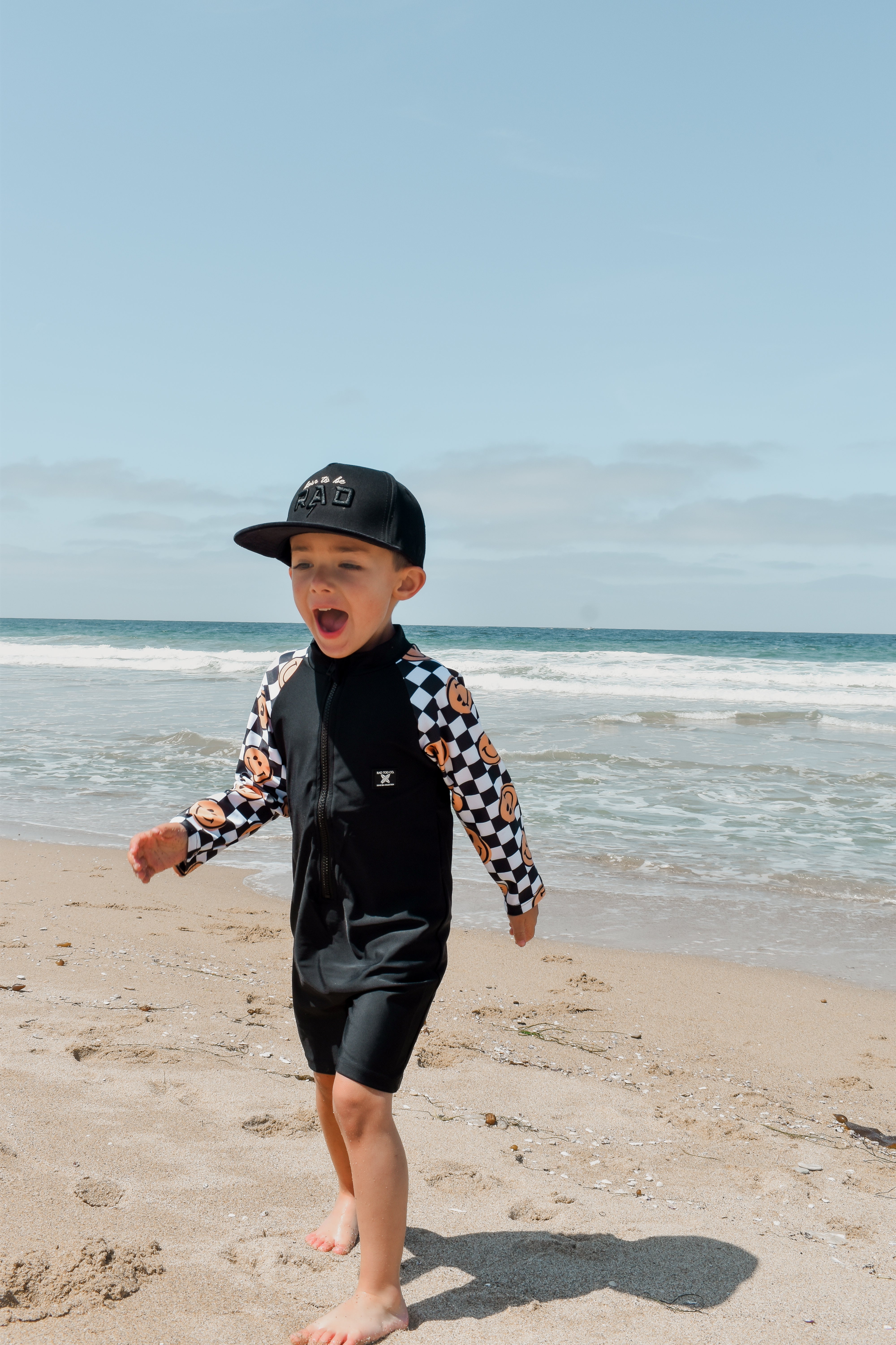 Young boy wearing a checker-patterned one-piece rashguard with smiley faces on the sleeves, standing on the beach near the shoreline