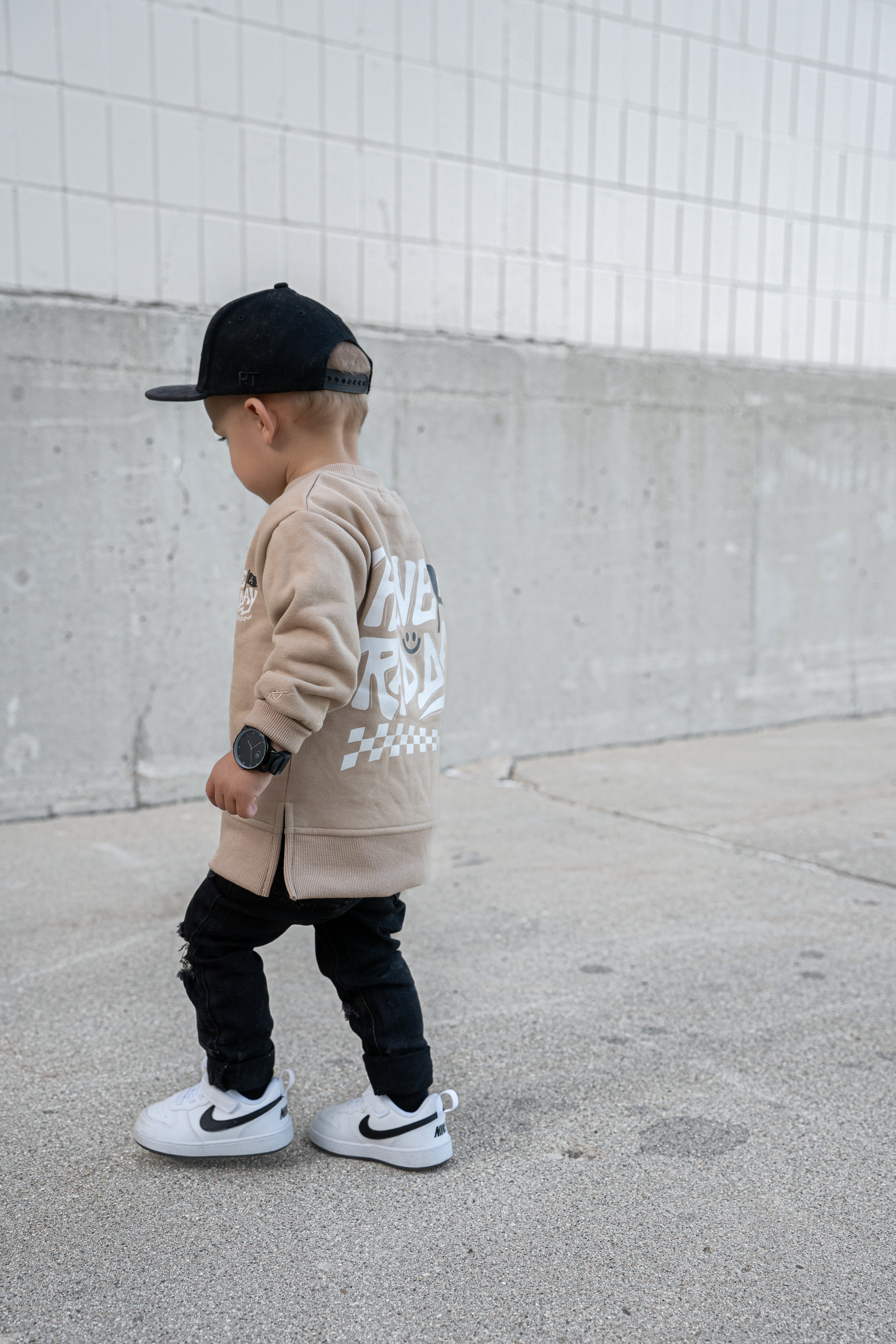 Child wearing a beige hoodie with text, black pants, and white sneakers against a concrete wall.