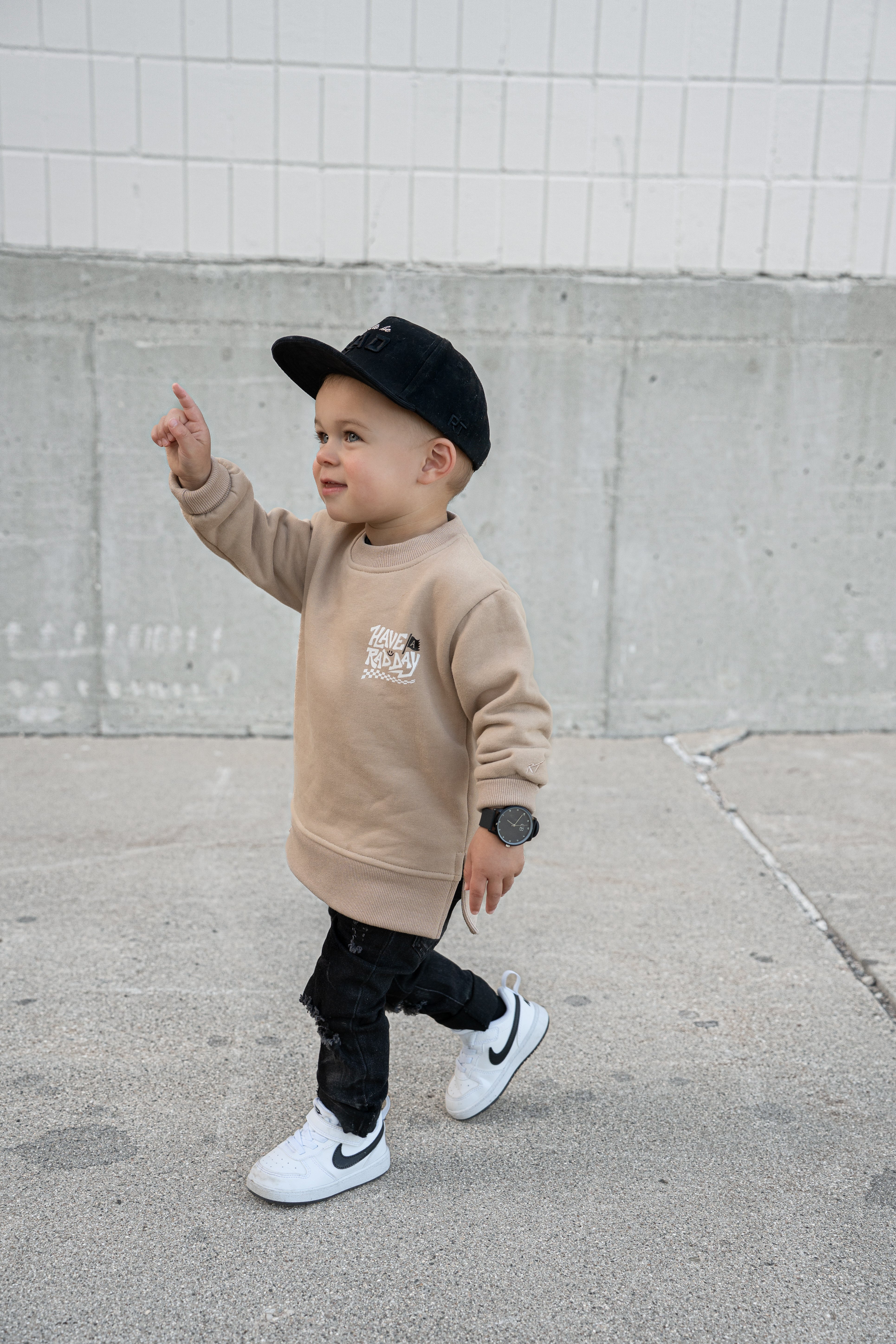 Child wearing a beige sweatshirt, black pants, and a black cap, standing on a sidewalk.