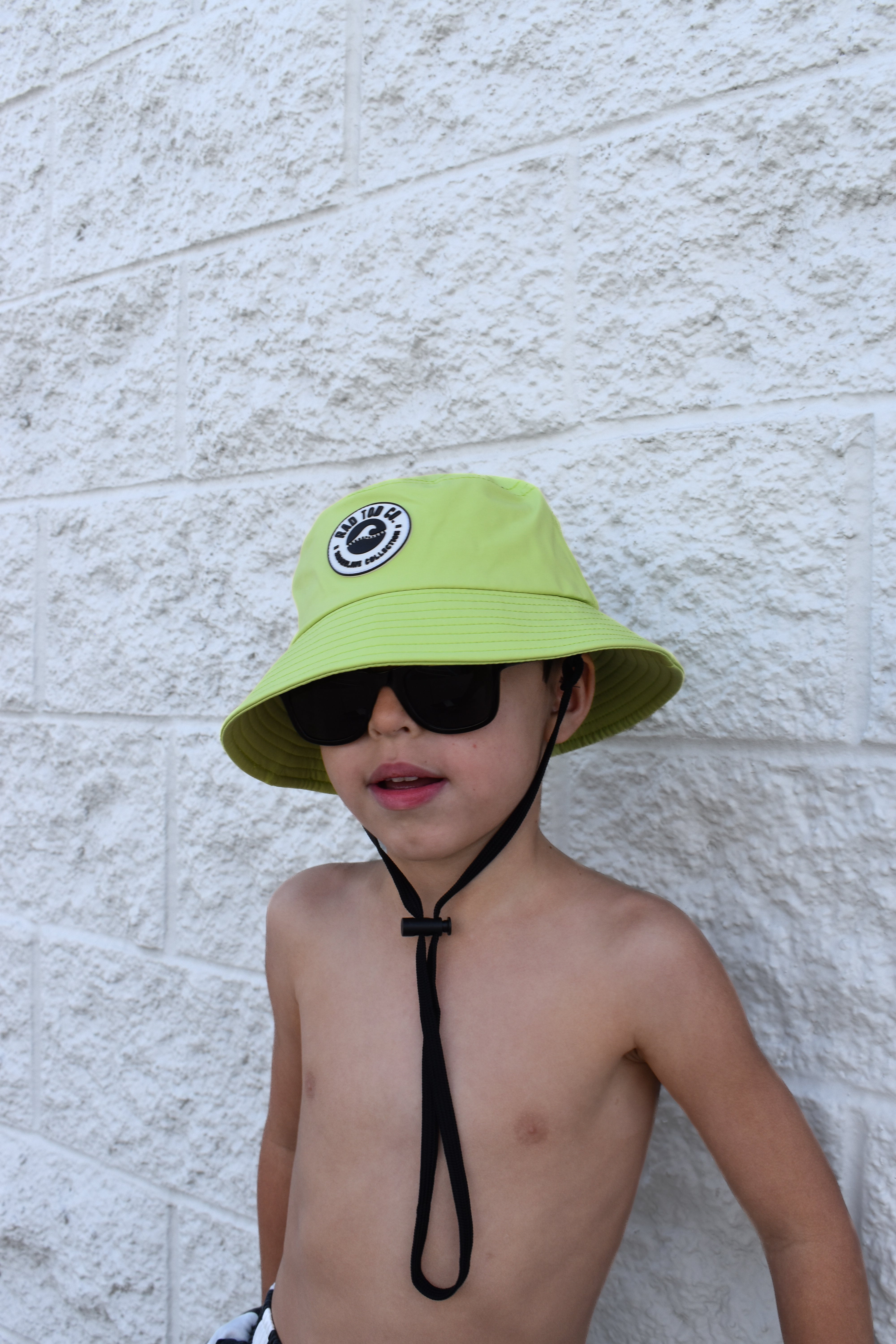 Child wearing a bright green bucket hat and sunglasses against a light stone wall.