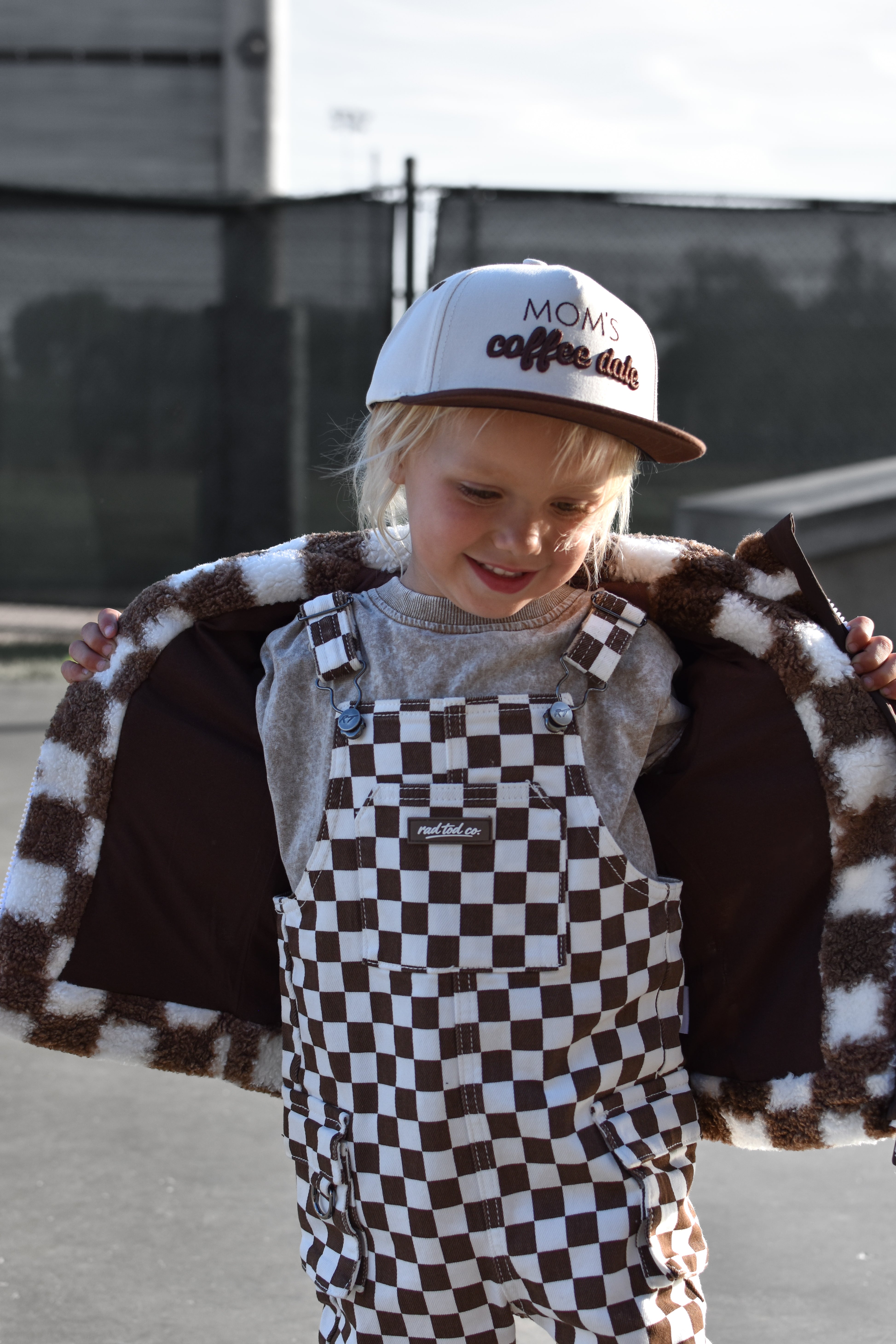 A young child wearing brown and white checkered overalls with a matching fuzzy checkered jacket draped open behind them, a gray shirt, and a white trucker hat that reads 'Moms coffee date'. The photo is taken outdoors on a paved surface with a fence and building in the background.