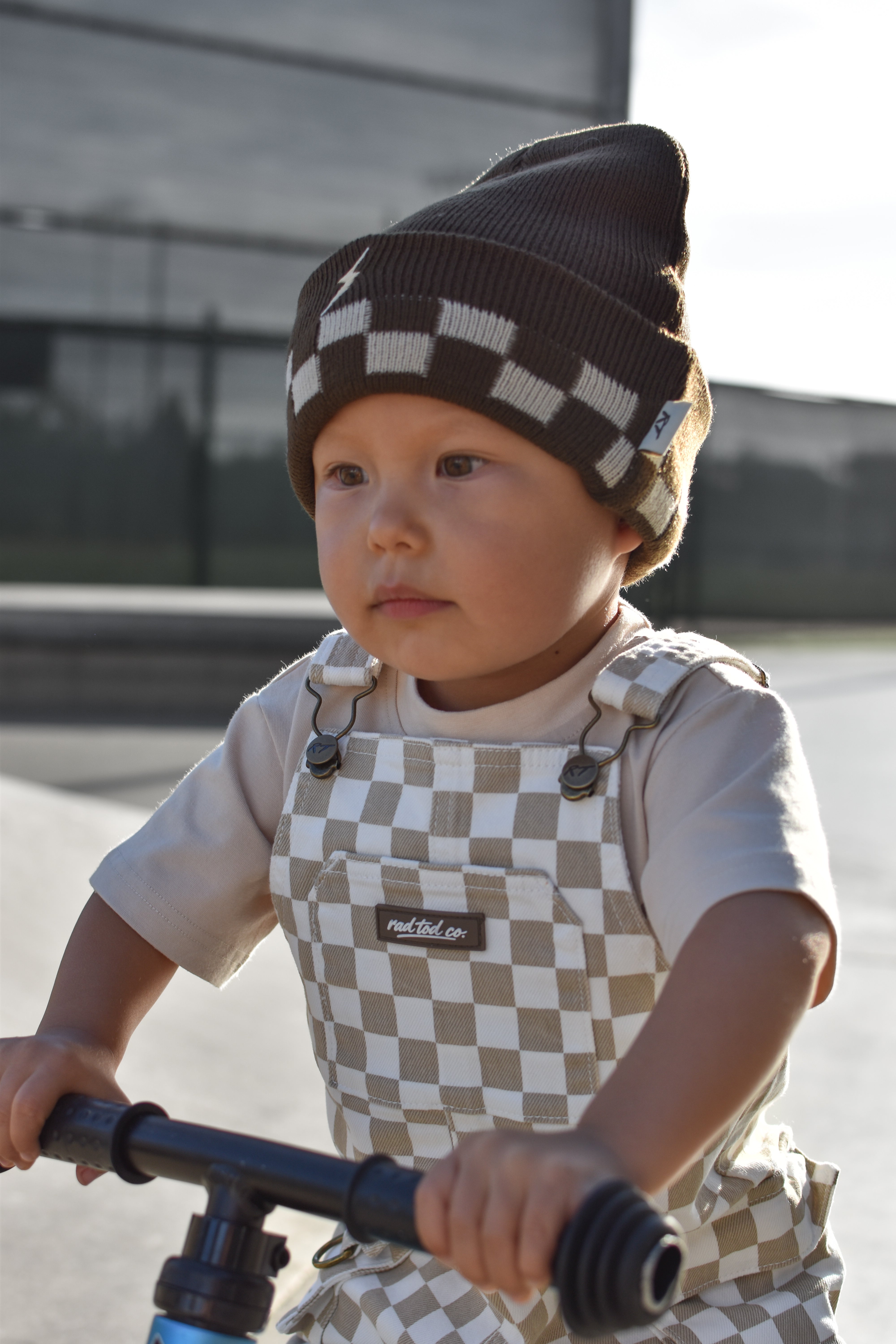 A toddler wearing a brown and beige checkered knit beanie and matching checkered overalls rides a blue bicycle outdoors.