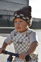 A toddler wearing a brown and beige checkered knit beanie and matching checkered overalls rides a blue bicycle outdoors.