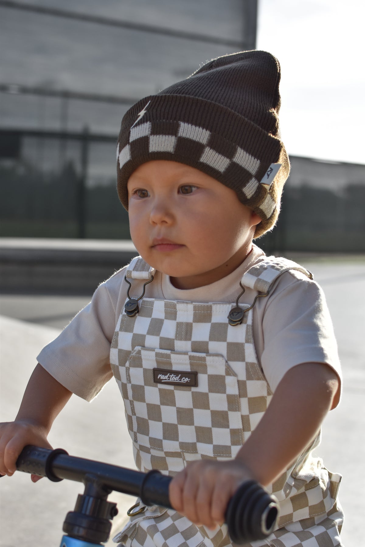 A toddler wearing a brown and beige checkered knit beanie and matching checkered overalls rides a blue bicycle outdoors.