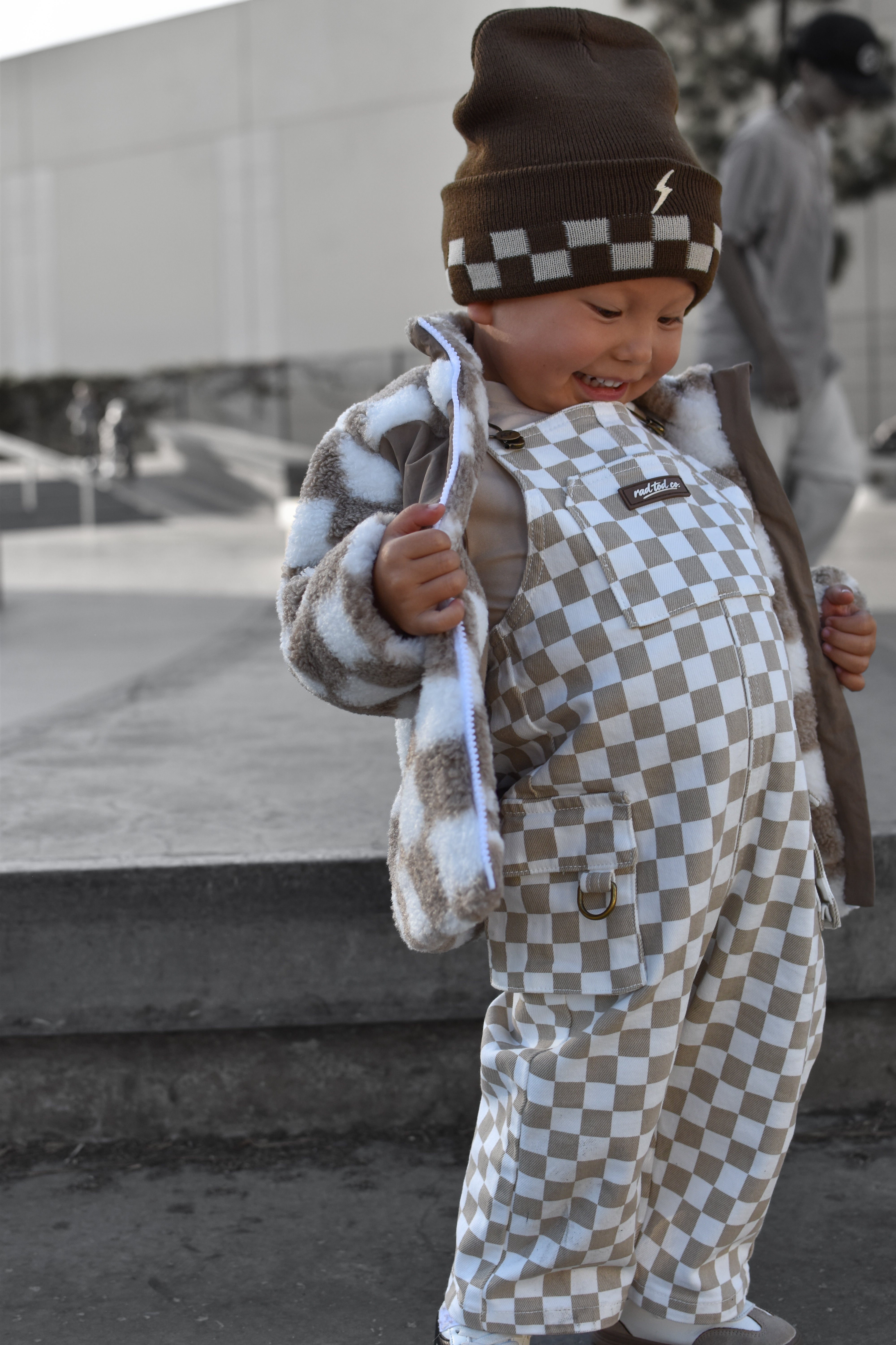 A toddler wearing a brown and beige checkered beanie, a brown and beige checkered overalls, and a brown and white checkered fleece jacket, standing outdoors in a city setting.