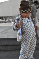 A toddler wearing a brown and beige checkered beanie, a brown and beige checkered overalls, and a brown and white checkered fleece jacket, standing outdoors in a city setting.