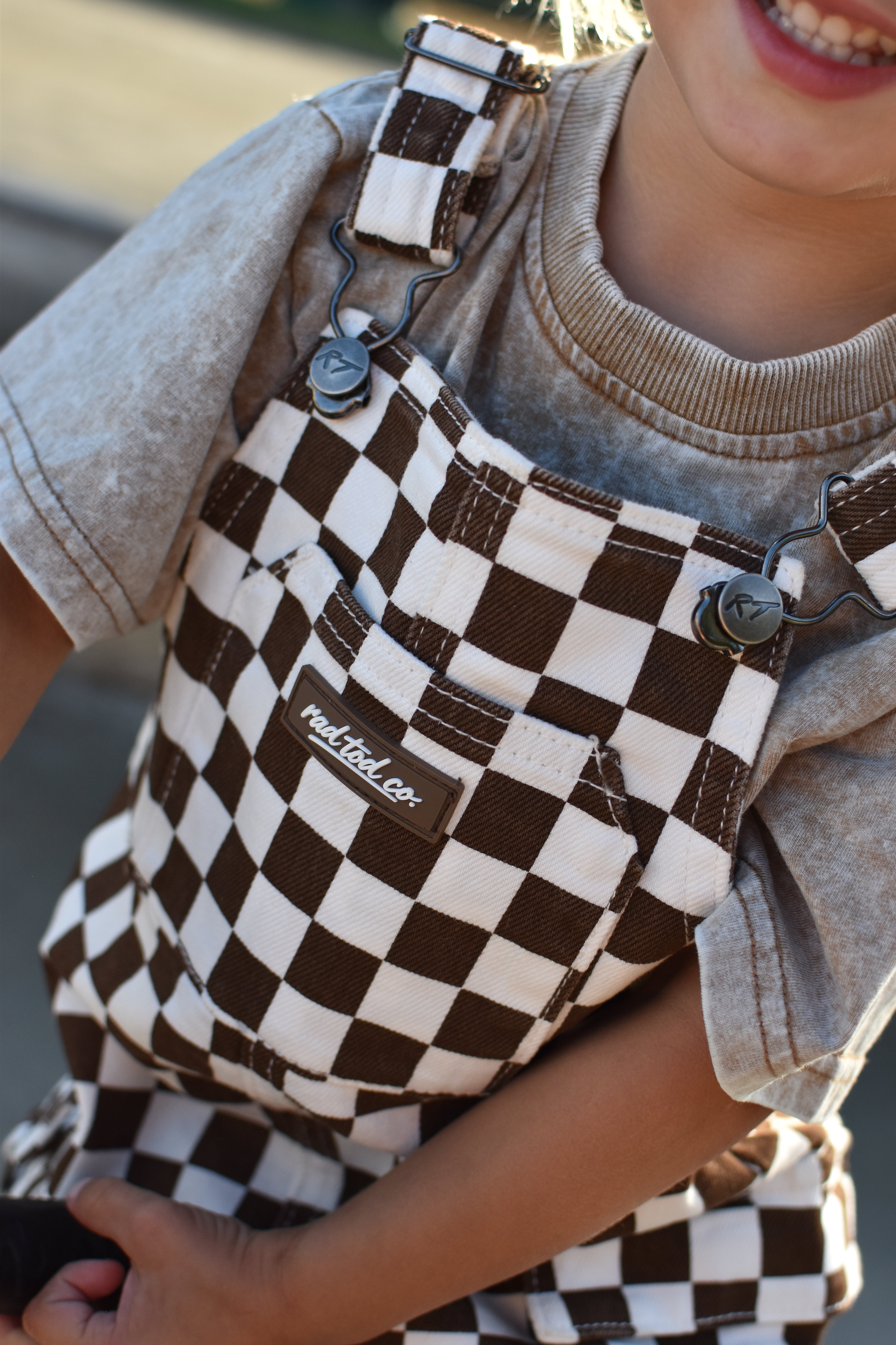 A close-up of a child wearing brown and cream checkered canvas overalls over a faded beige t-shirt. The brand name Rad Tod Co.