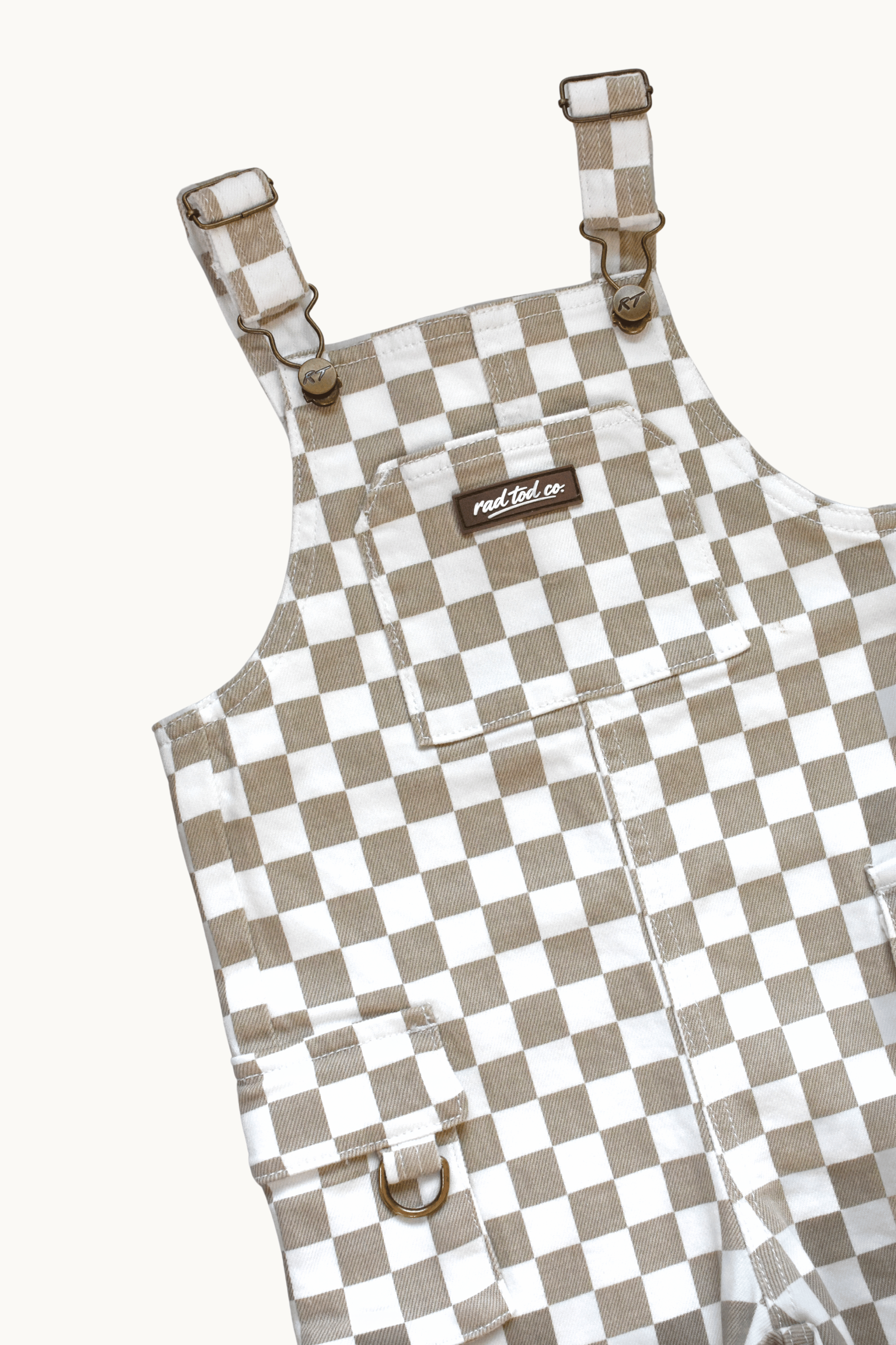 A close-up of a pair of checkered overalls in a beige and white gingham pattern, featuring adjustable shoulder straps with metal buckles, a front chest pocket, and a small brown brand label reading rad tod co,