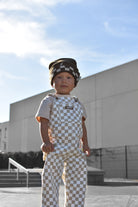 A toddler standing outdoors in front of a modern building, wearing a matching beige and white checkerboard outfit with a small hat.