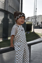 A toddler standing outdoors on a concrete path, wearing brown and beige checkered overalls over a light beige t-shirt and a matching checkered beanie.
