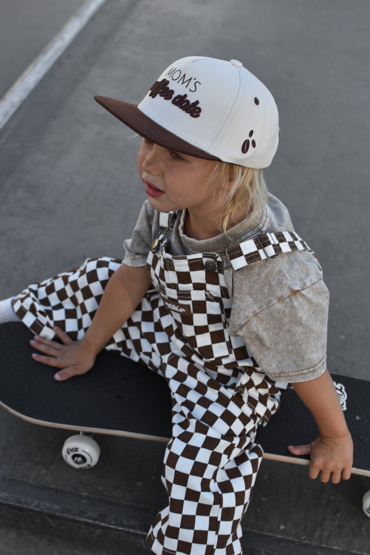 A child wearing brown and white checkered overalls and a matching baseball cap sits on a skateboard on an asphalt surface.