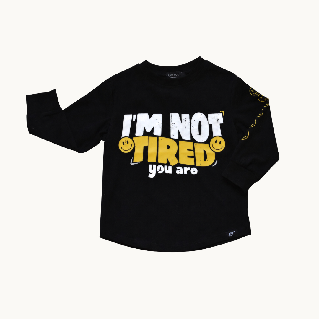 A black long-sleeve t-shirt with a graphic print on the front reading 'I'M NOT TIRED you are' in white and yellow, accompanied by smiley faces, shown on a light cream background.
