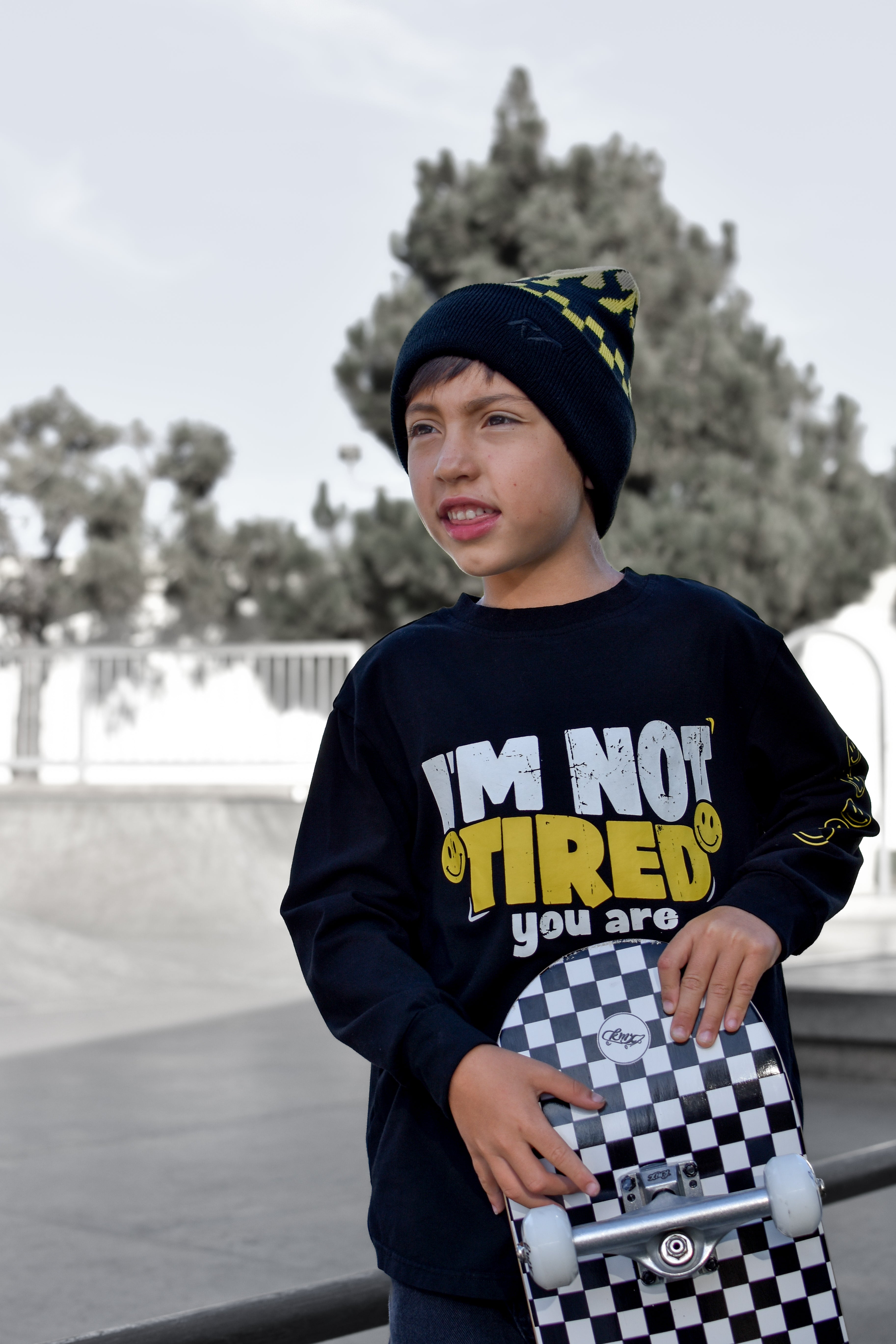 A young boy wearing a black beanie with yellow accents and a black graphic sweatshirt that reads 'I'M NOT TIRED you are' holds a checkered skateboard in an outdoor skatepark setting.