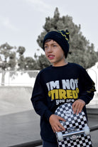 A young boy wearing a black beanie with yellow accents and a black graphic sweatshirt that reads 'I'M NOT TIRED you are' holds a checkered skateboard in an outdoor skatepark setting.