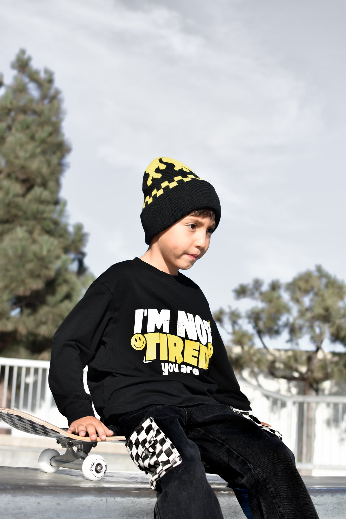 A child wearing a black graphic sweatshirt with yellow and white text, a black beanie with yellow checkers, and black pants with a checkered pattern, sitting on a skateboard outdoors.