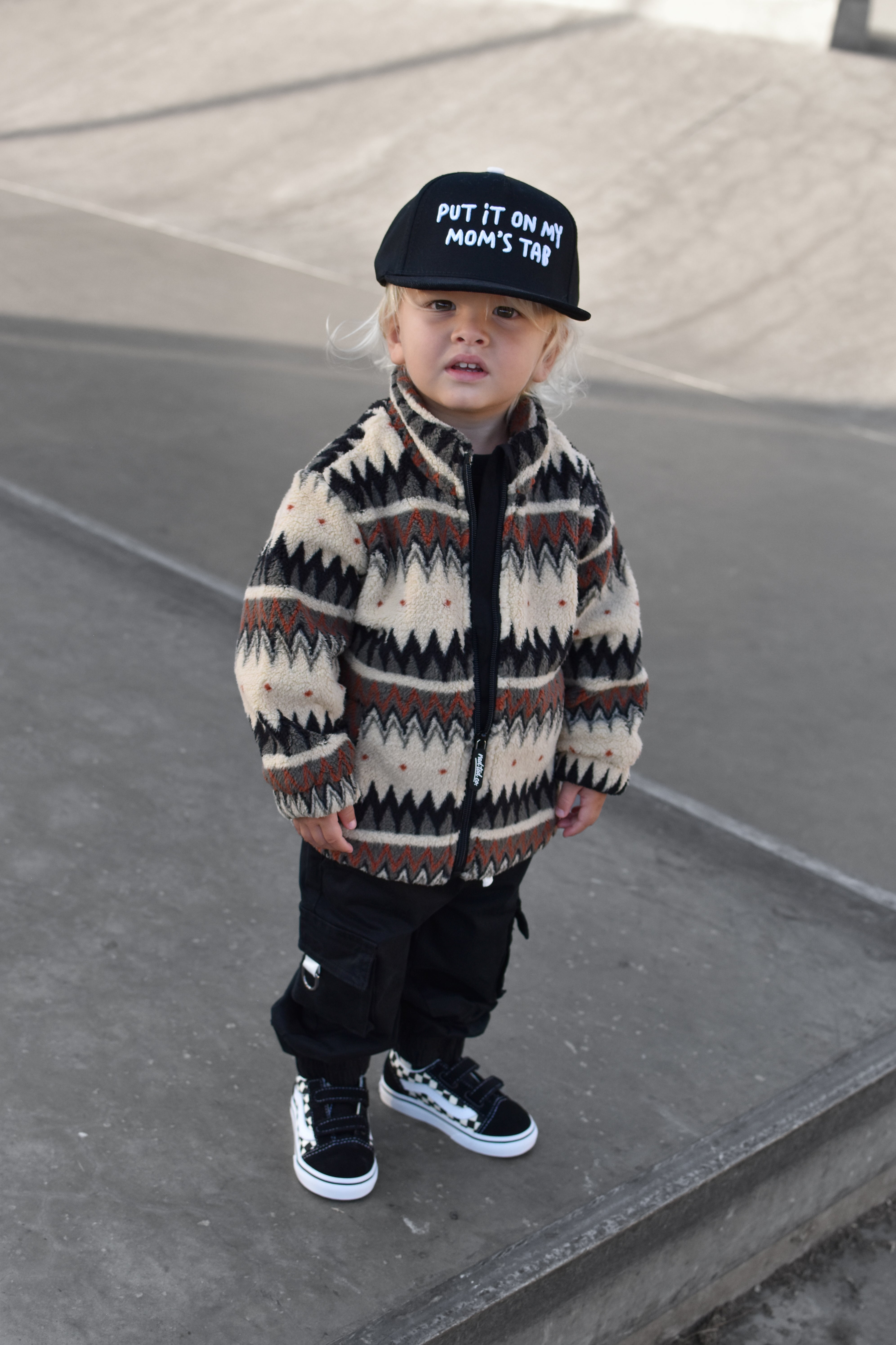 A toddler standing outdoors on concrete steps, wearing a cream and brown geometric patterned zip-up jacket, black pants, black sneakers, and a black cap with white text that reads 'PUT IT ON MY MOM'S TAB'.