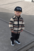 A toddler standing outdoors on concrete steps, wearing a cream and brown geometric patterned zip-up jacket, black pants, black sneakers, and a black cap with white text that reads 'PUT IT ON MY MOM'S TAB'.