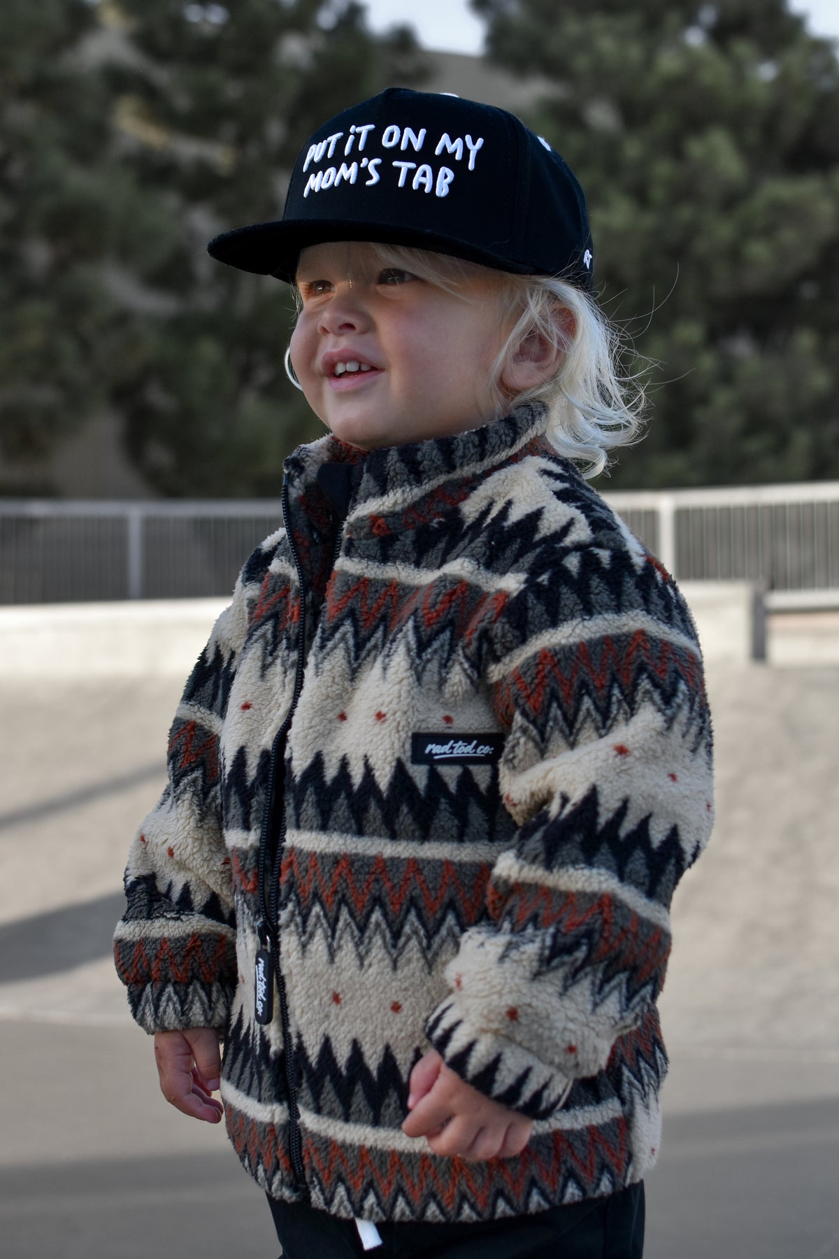 A toddler wearing a cream, black, and rust-colored geometric patterned fleece jacket and a black baseball cap that says put it on my moms tab, standing outdoors at a skatepark.