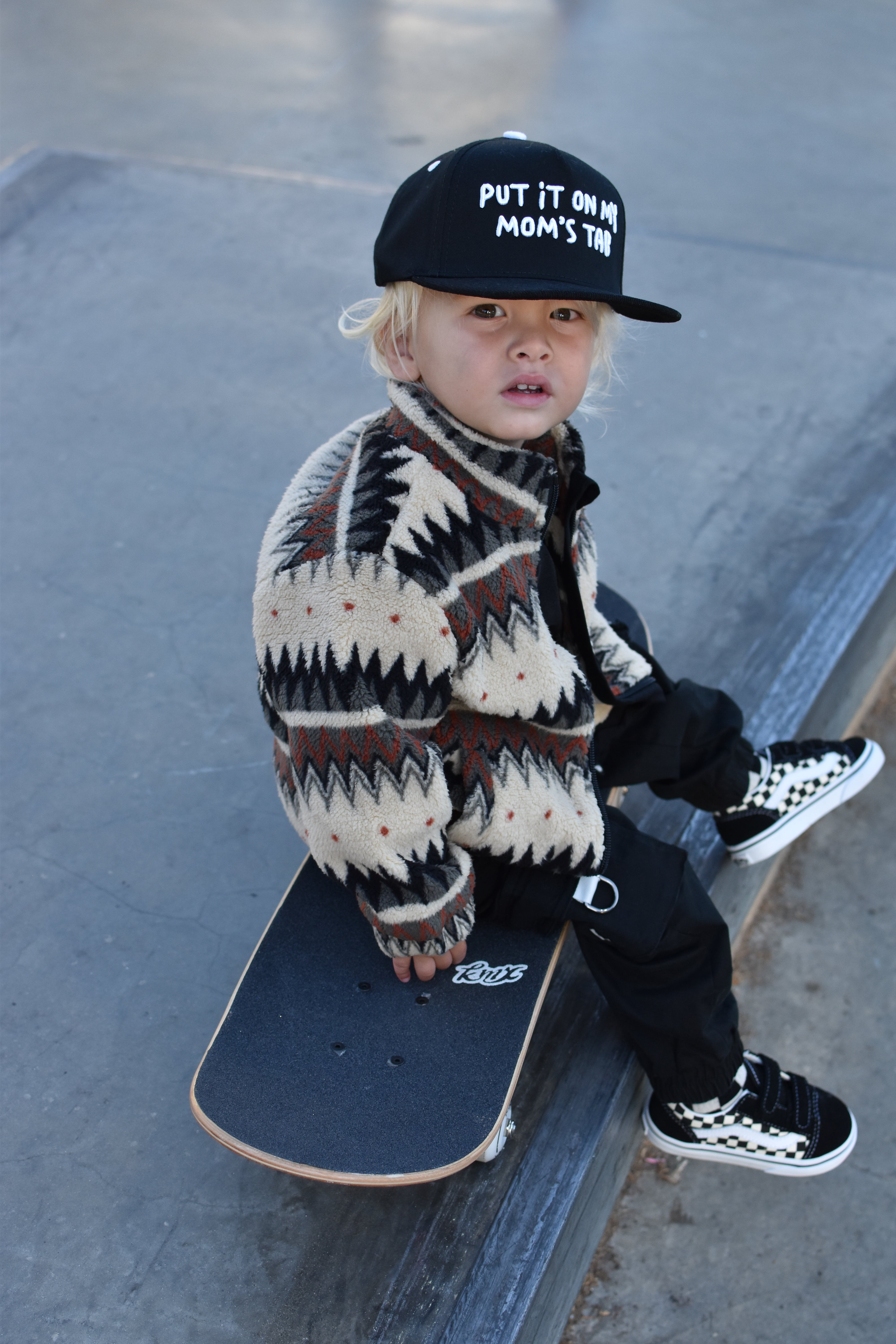 A toddler sits on a skateboard on a concrete ramp, wearing a black cap with white text, a cream and black patterned knit sweater, black pants, and black-and-white checkered sneakers.
