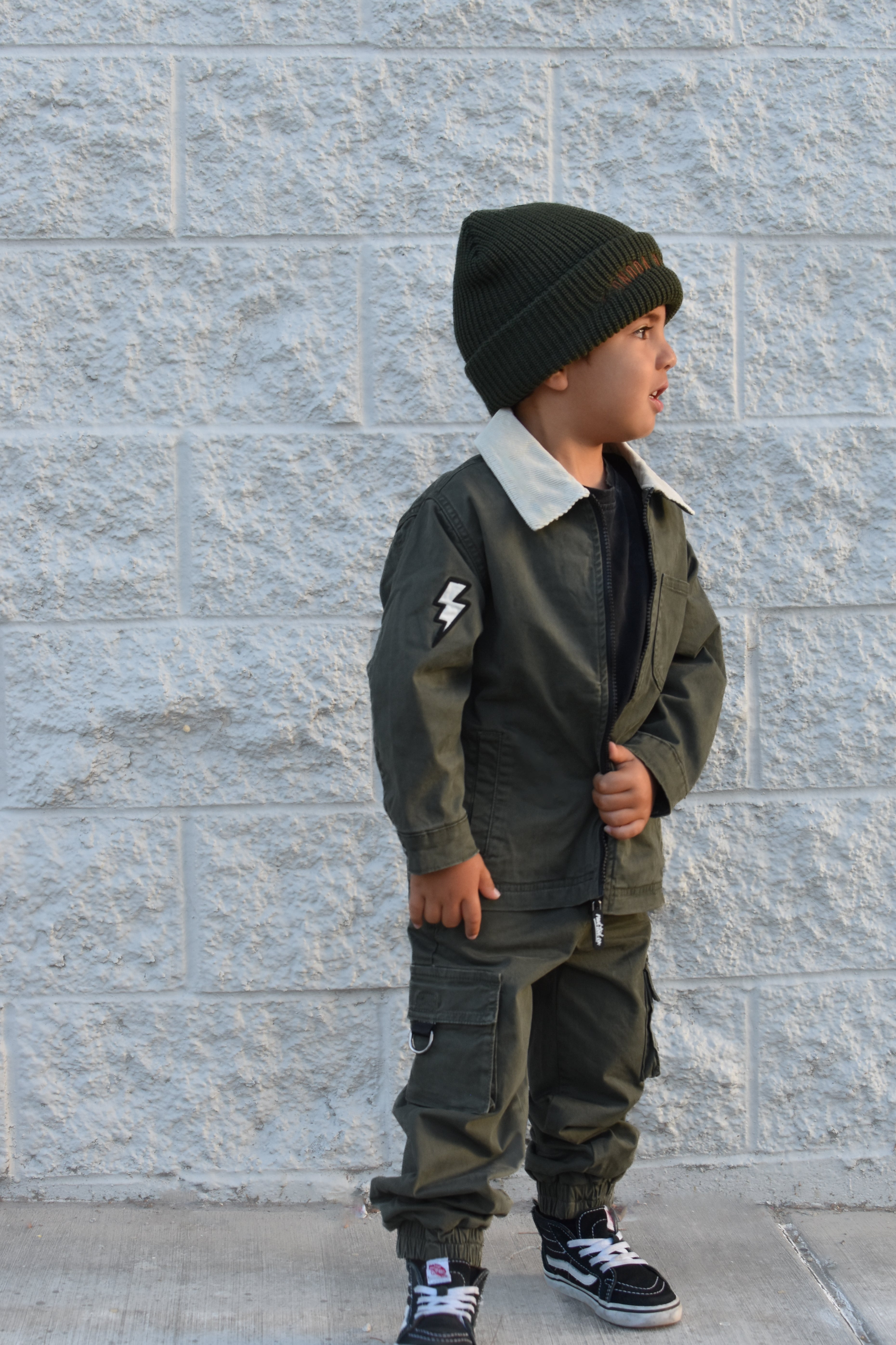 A young child wearing an olive green two-piece jacket and cargo pants set with a white collar, a black beanie, and black sneakers, standing against a light gray textured brick wall.