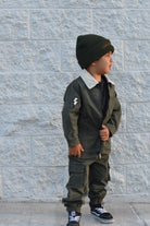 A young child wearing an olive green two-piece jacket and cargo pants set with a white collar, a black beanie, and black sneakers, standing against a light gray textured brick wall.